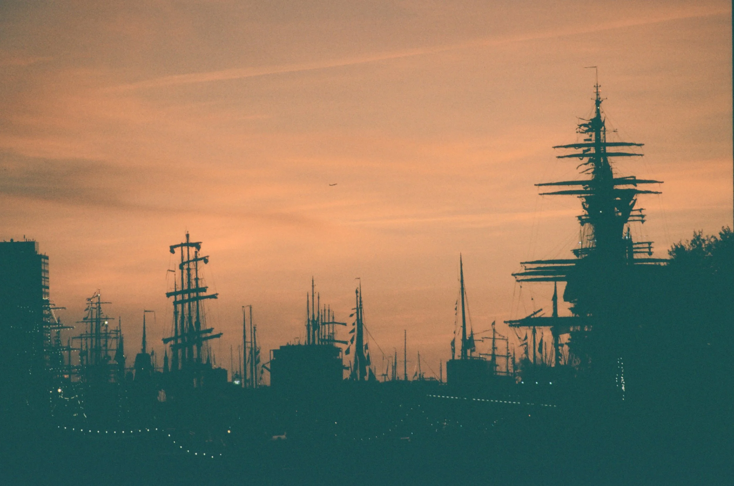 Silhouettes of tall ships with masts and rigging against a pink and orange sunset sky, near the city skyline with some buildings and trees.