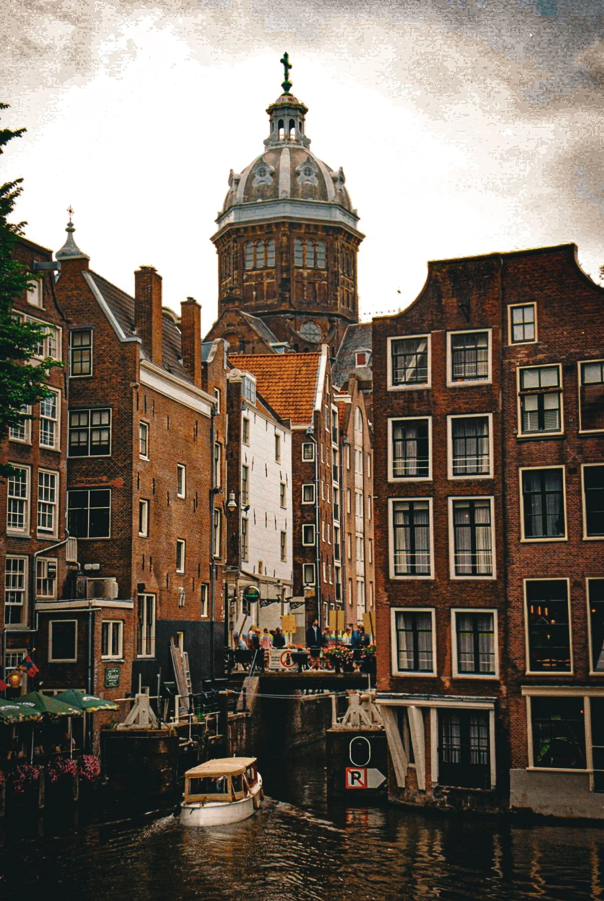 A canal in a European city with historic brick buildings, a boat on the water, and a large domed tower in the background.