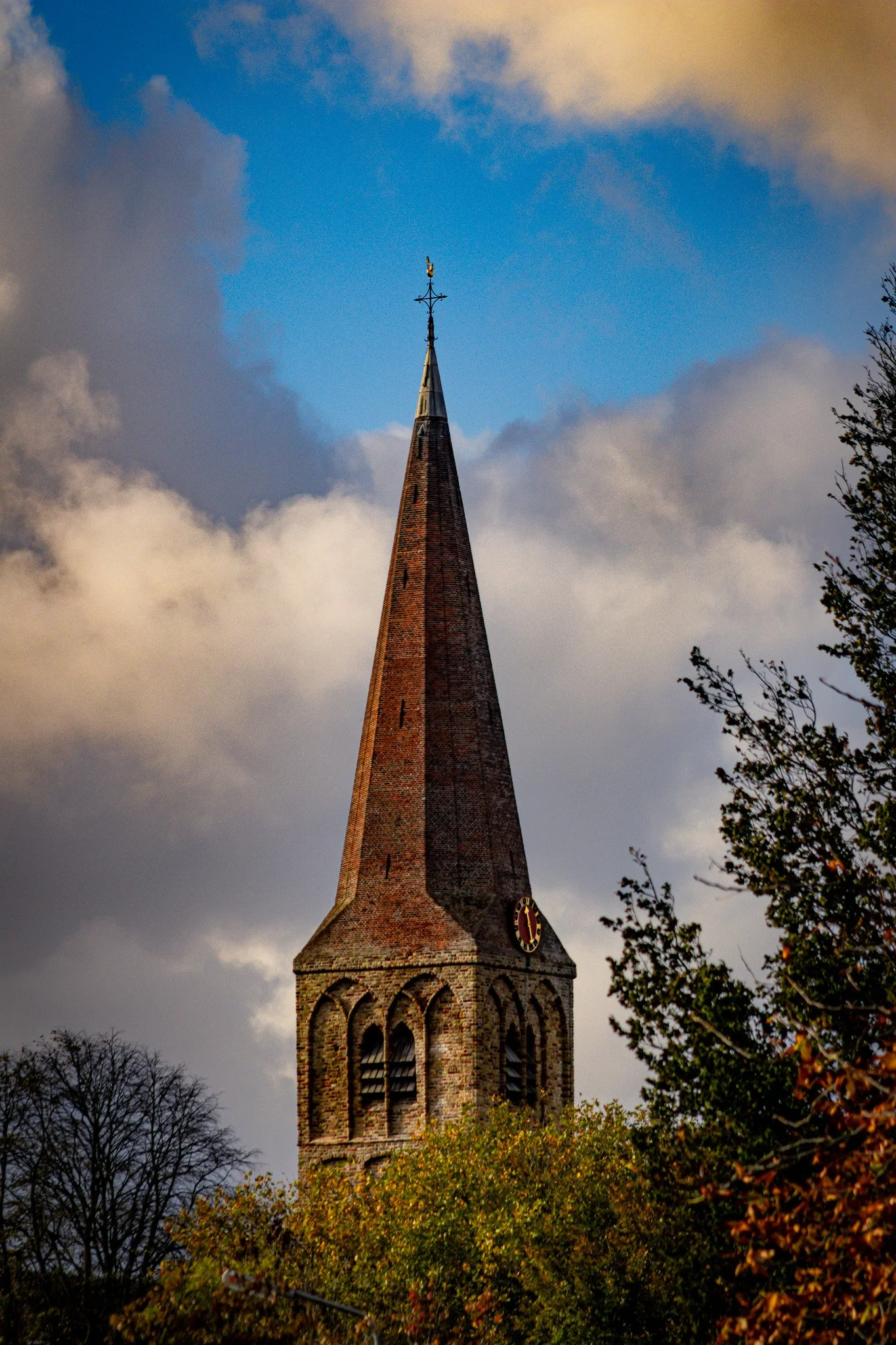 A tall brick church steeple with a clock on its side, surrounded by trees with autumn foliage under cloudy sky.