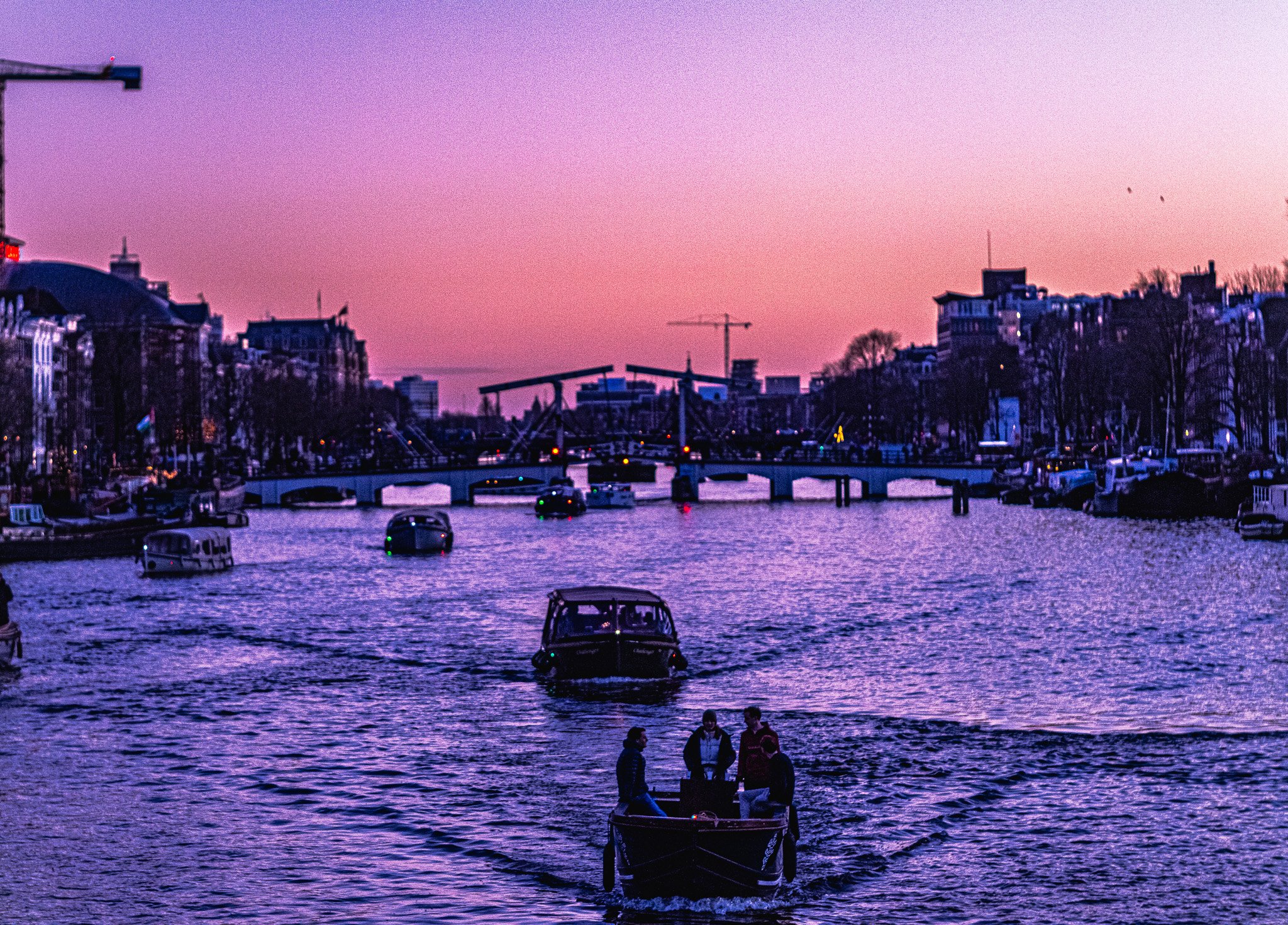 A boat with three people on a river during sunset, with buildings, bridges, and boats in the background.