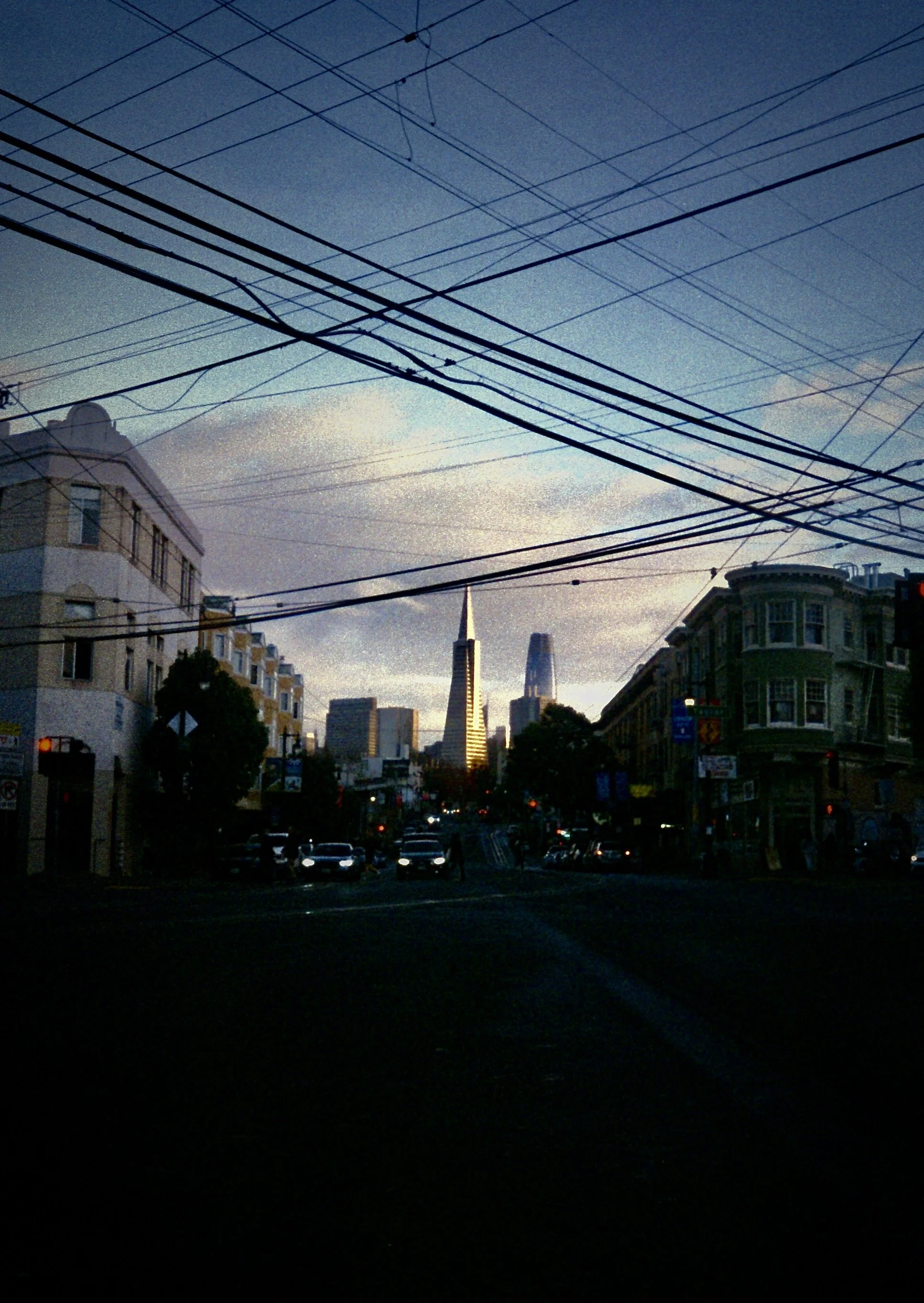 City street view with overhead utility wires, buildings on both sides, and a skyline featuring the Salesforce Tower and the Transamerica Pyramid at sunset.