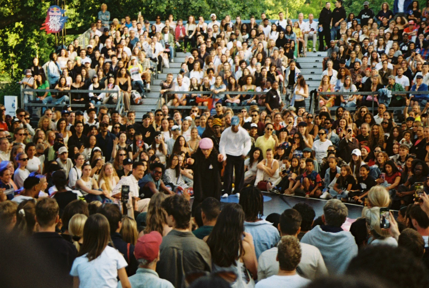 Large outdoor gathering with audience seated on bleachers and ground, watching a street performance on a circular stage, with two performers in center, surrounded by trees and clear sky.