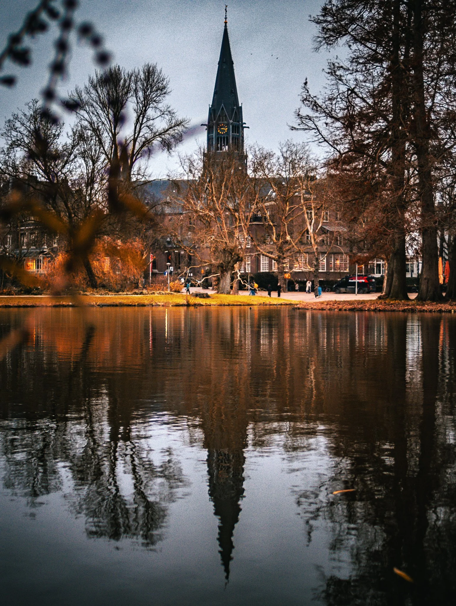 A church with a tall spire reflected in a body of water surrounded by trees and a park with people walking.