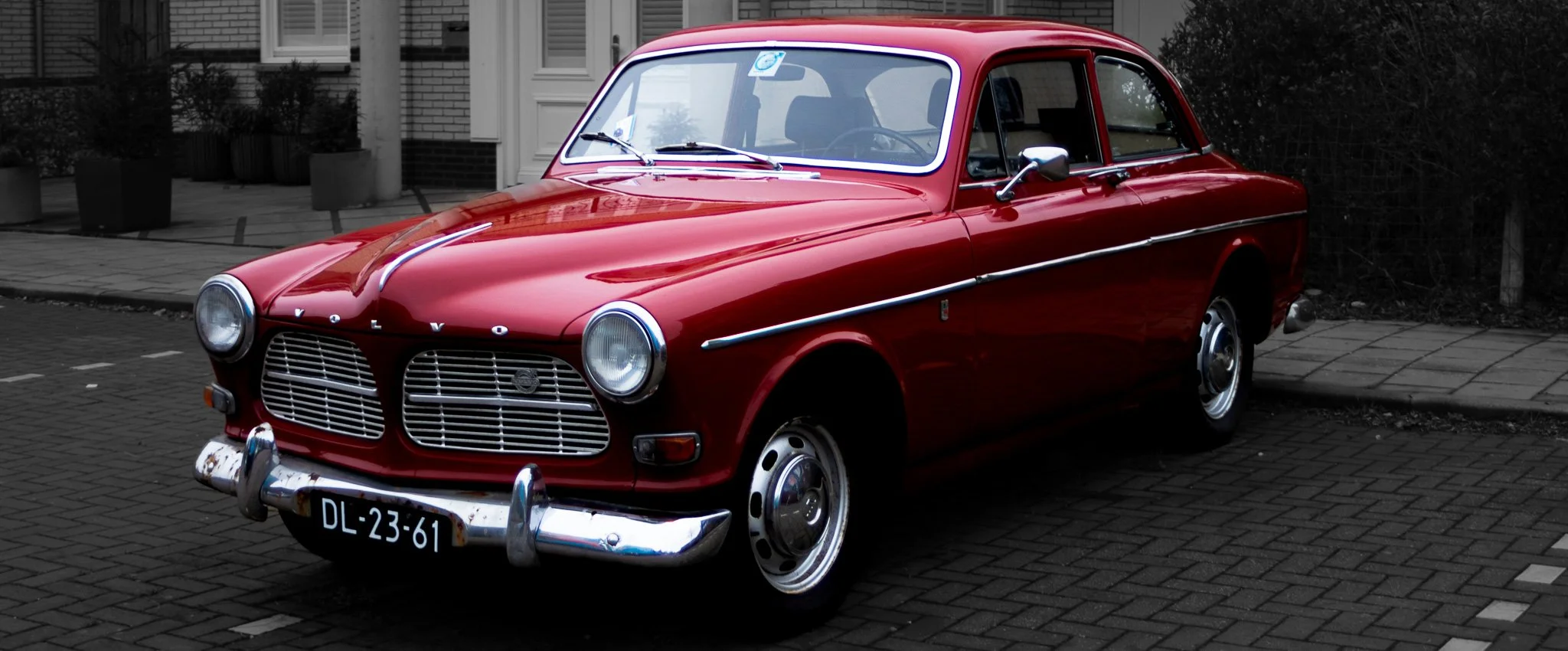 Red vintage Volvo car parked on a city street with brick pavement, with a black and white background.