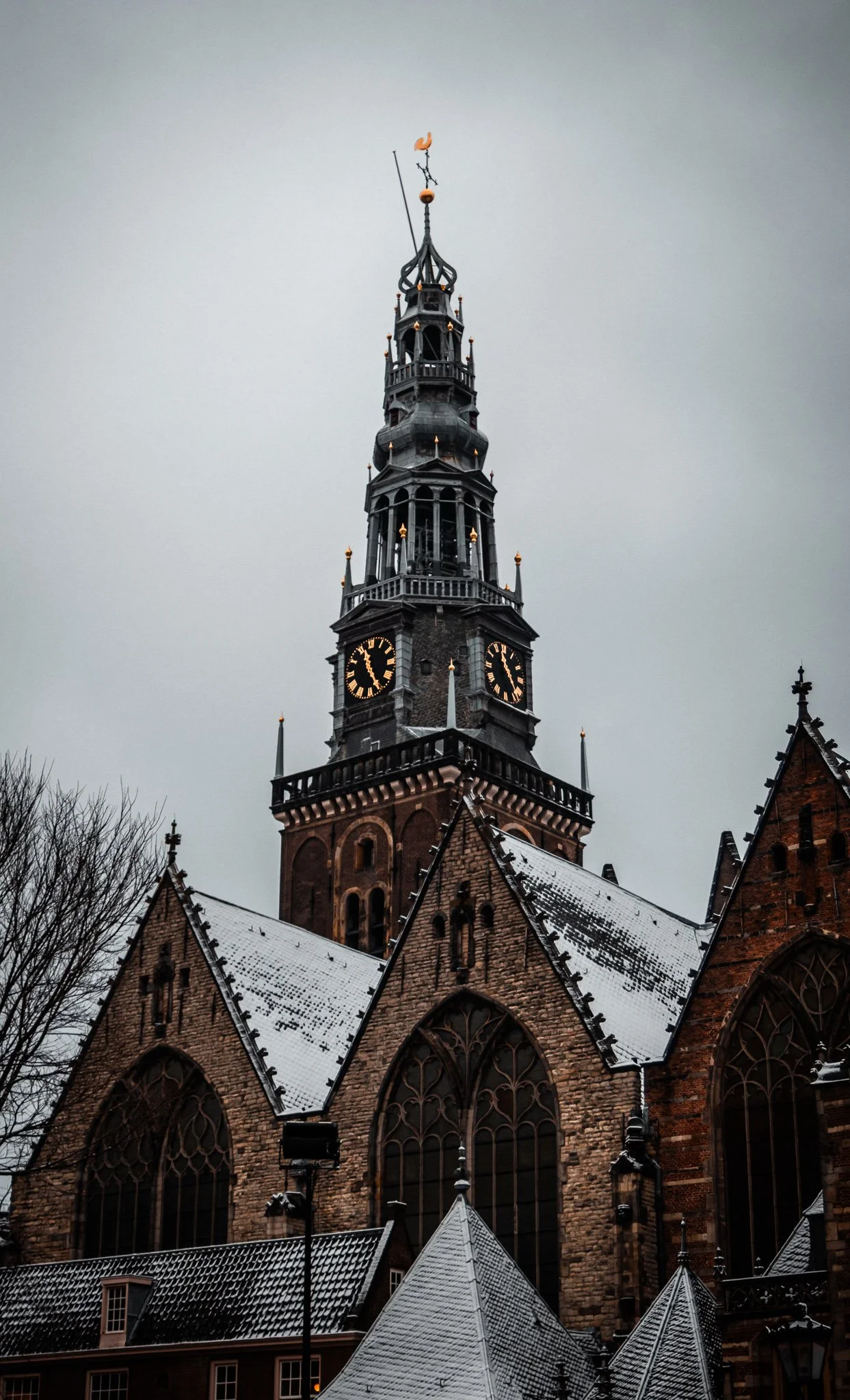 A tall, gothic-style church steeple with a clock, topped with a weather vane, against a gray sky and snow-covered roof.