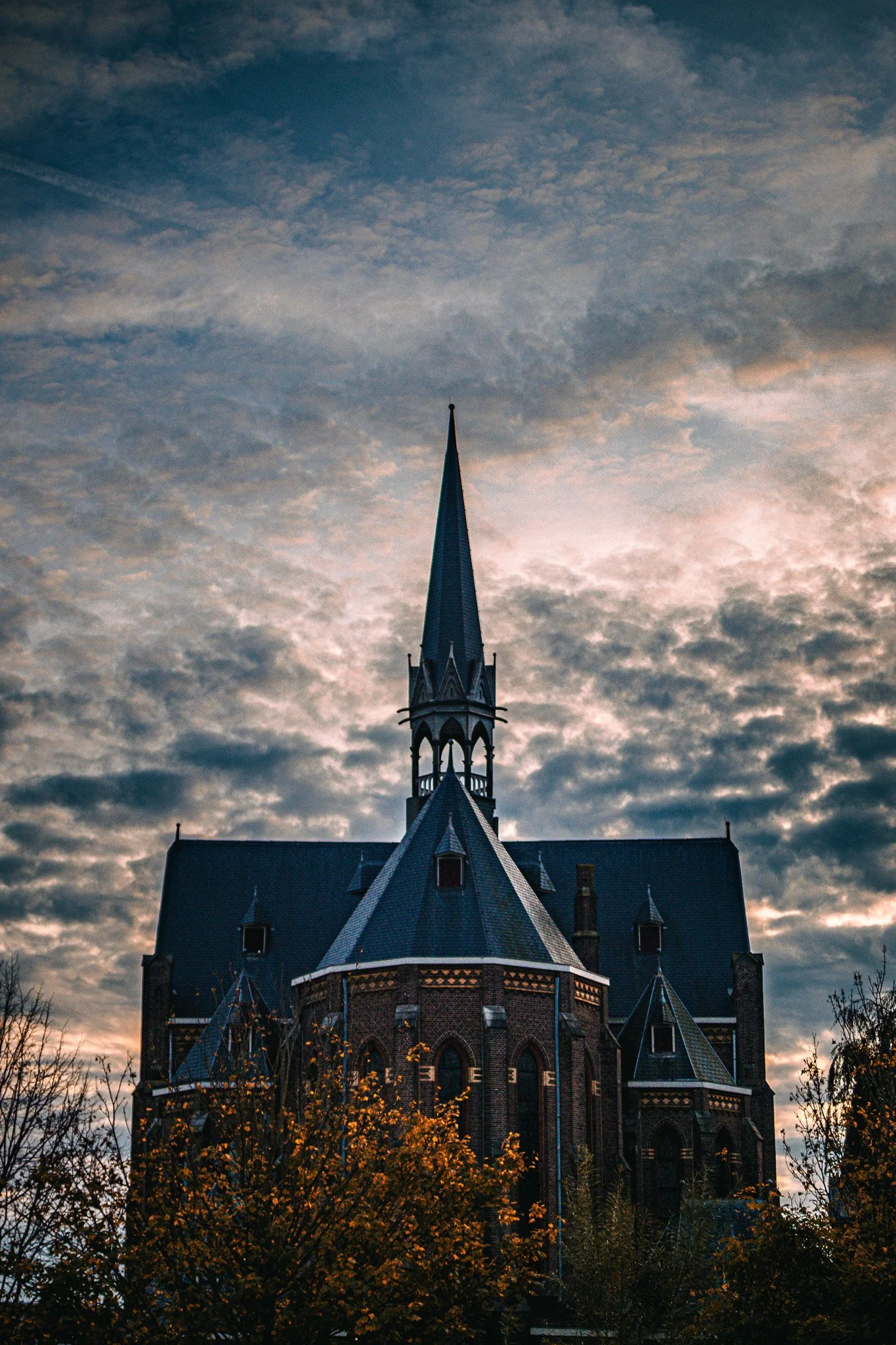 A tall, pointed church steeple rises against a cloudy sky at sunset, with trees in the foreground.