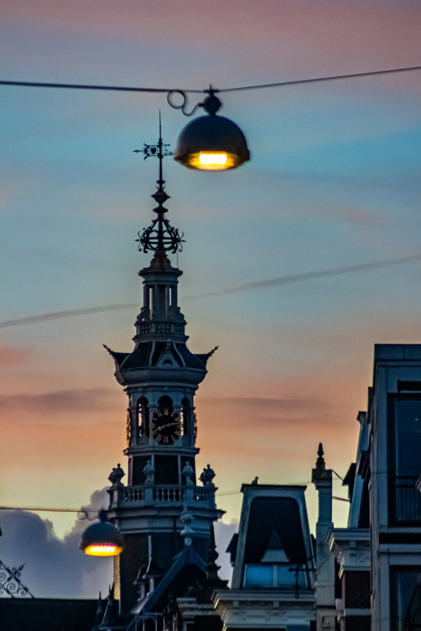 A cityscape at sunset featuring a historic clock tower and two illuminated street lamps suspended overhead.
