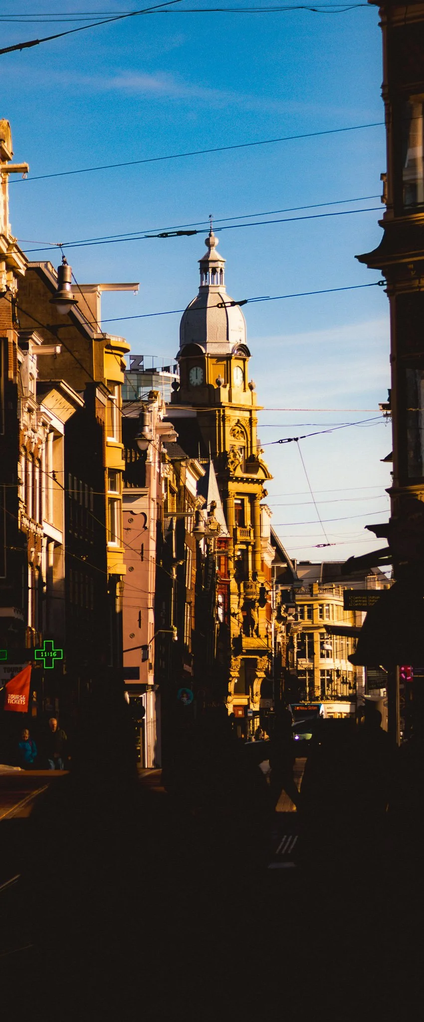 City street with historic buildings, a clock tower in the background, and people walking, during sunset.
