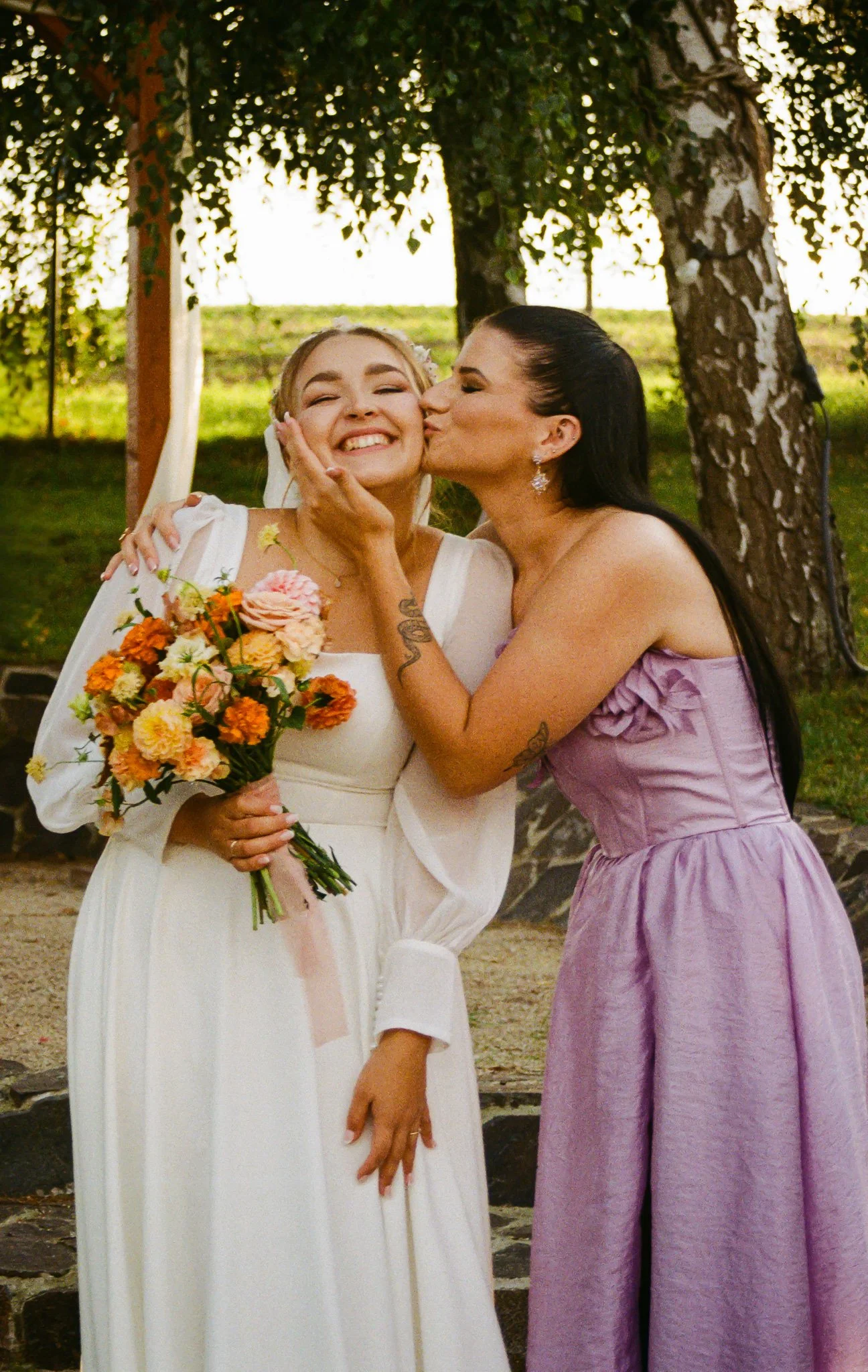 A wedding scene outdoors with a bride in a white dress holding a bouquet of flowers, kissing a woman in a lavender dress on the cheek, both smiling happily.