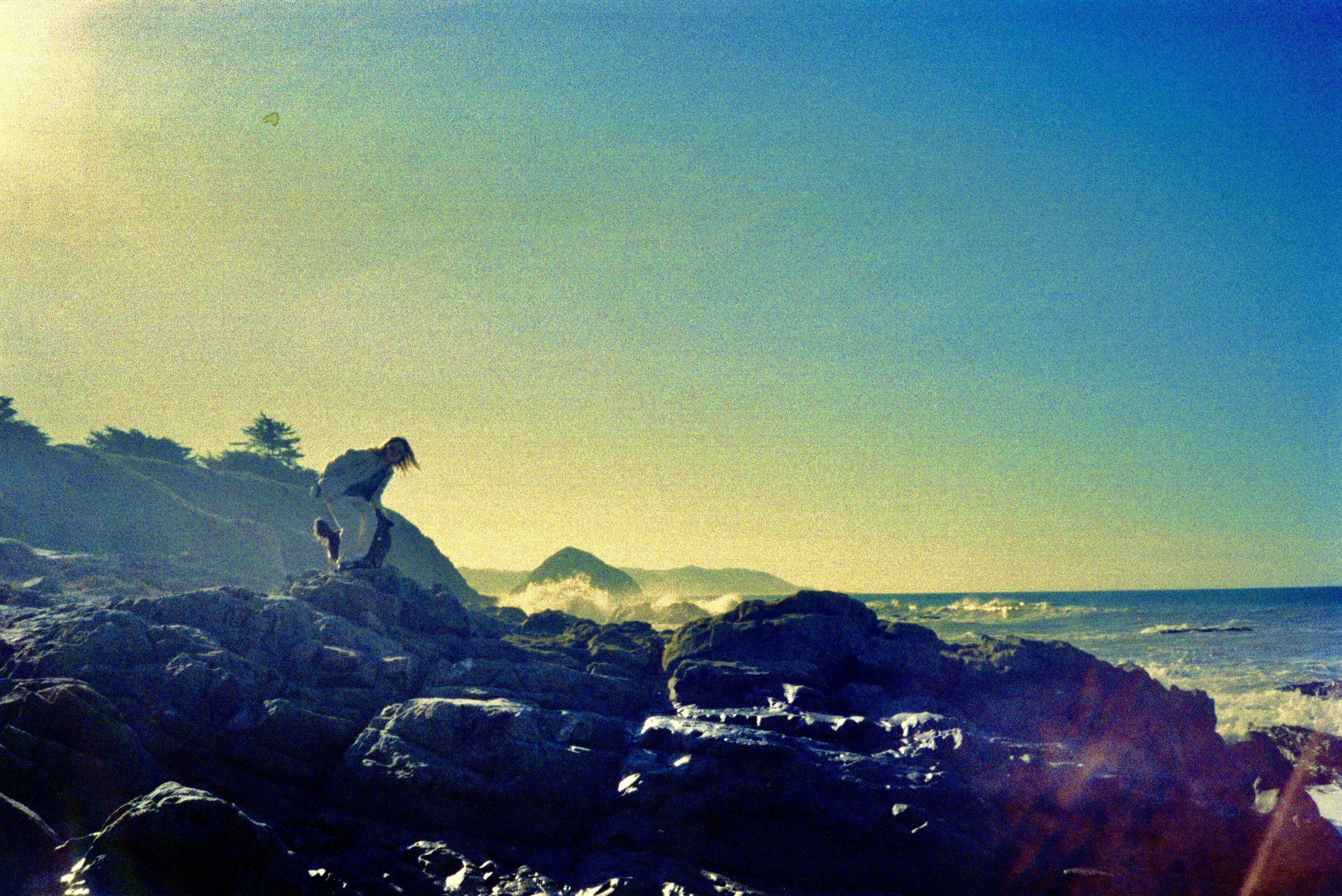 Person crouching on rocks near the ocean, with waves crashing and hills in the background under a clear sky.