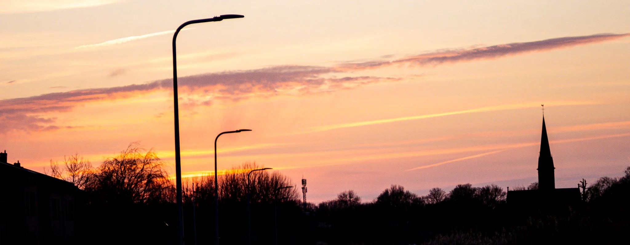 Sunset sky over a town silhouette with leafless trees, a church steeple, and streetlights.
