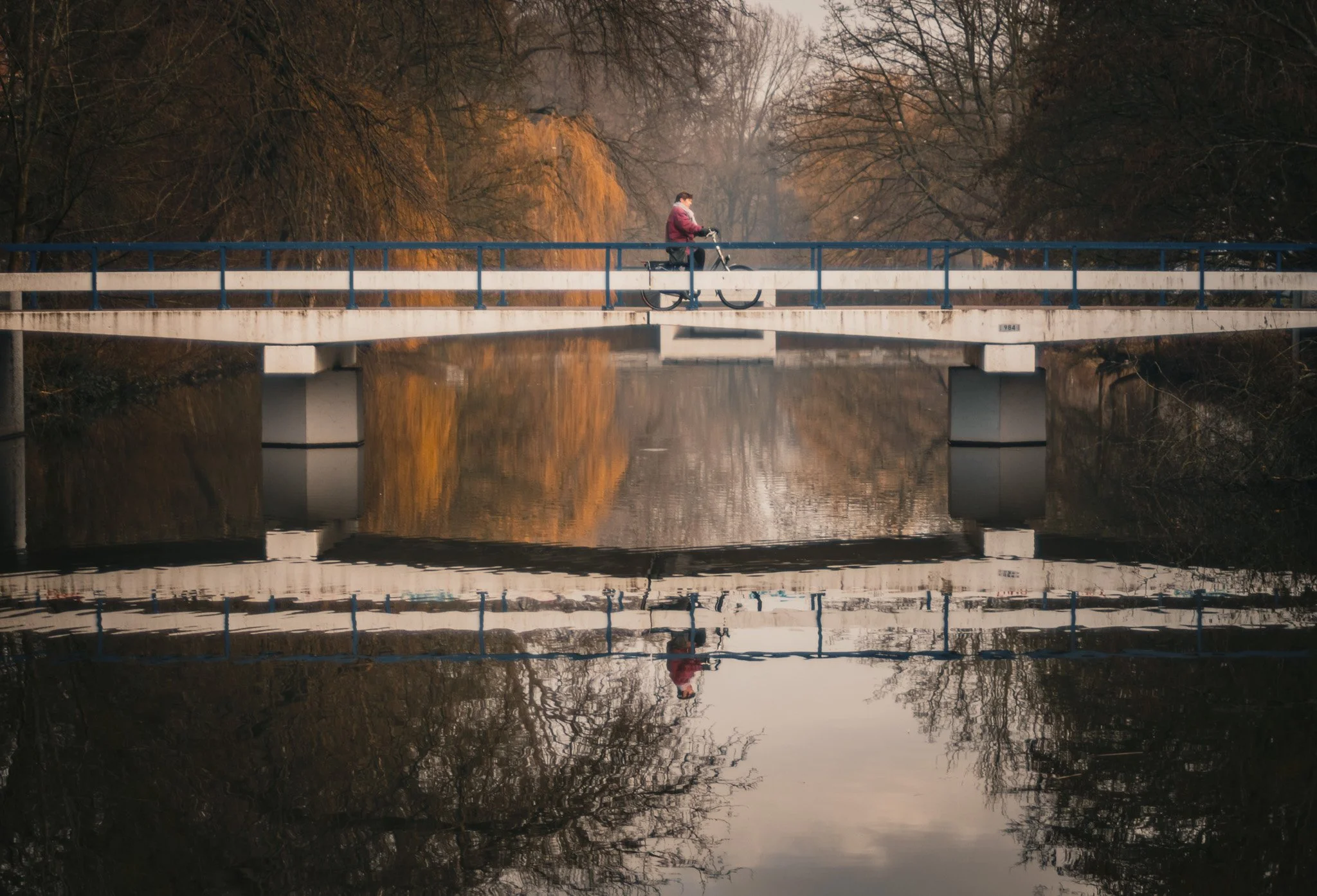 A person riding a bicycle across a bridge over a calm river, reflecting the bridge and surrounding trees.