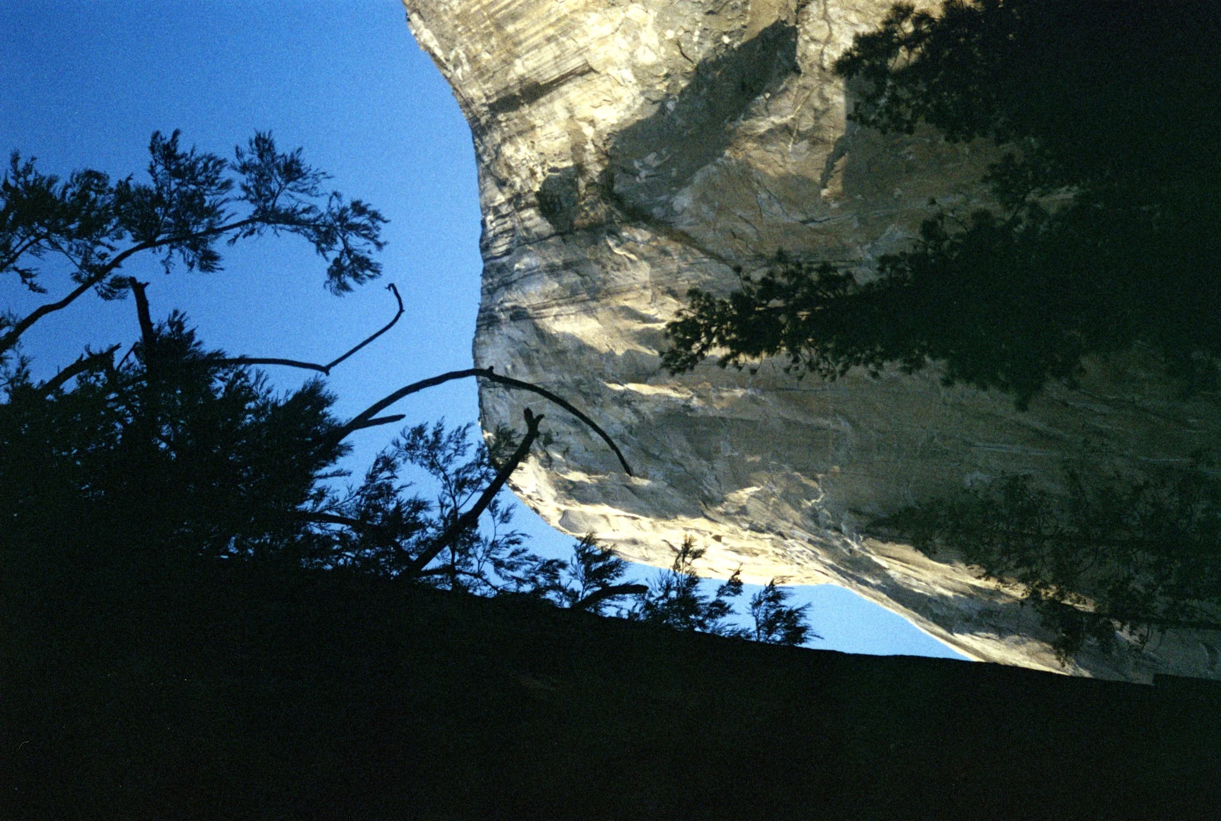 A mountain with a steep, rocky face illuminated by sunlight, with dark silhouettes of trees in the foreground and a clear blue sky.