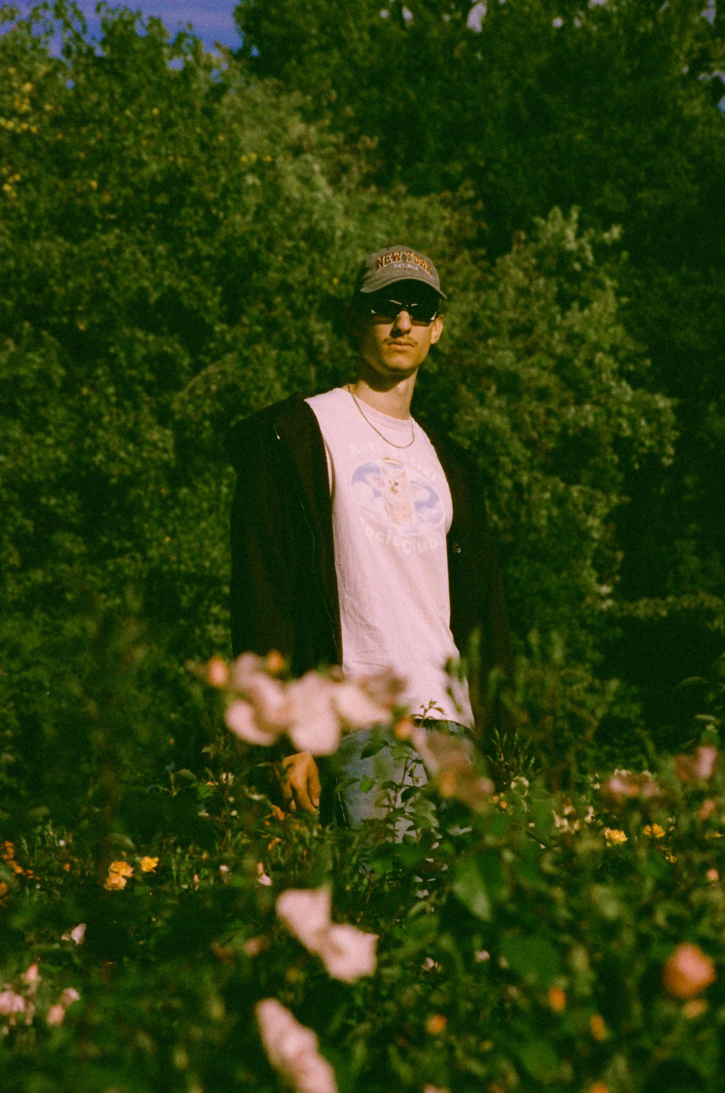 A young man stands outdoors among green bushes and trees, wearing sunglasses, a gray cap with 'New York' on it, a white T-shirt with a graphic design, a black jacket, and a gold chain.