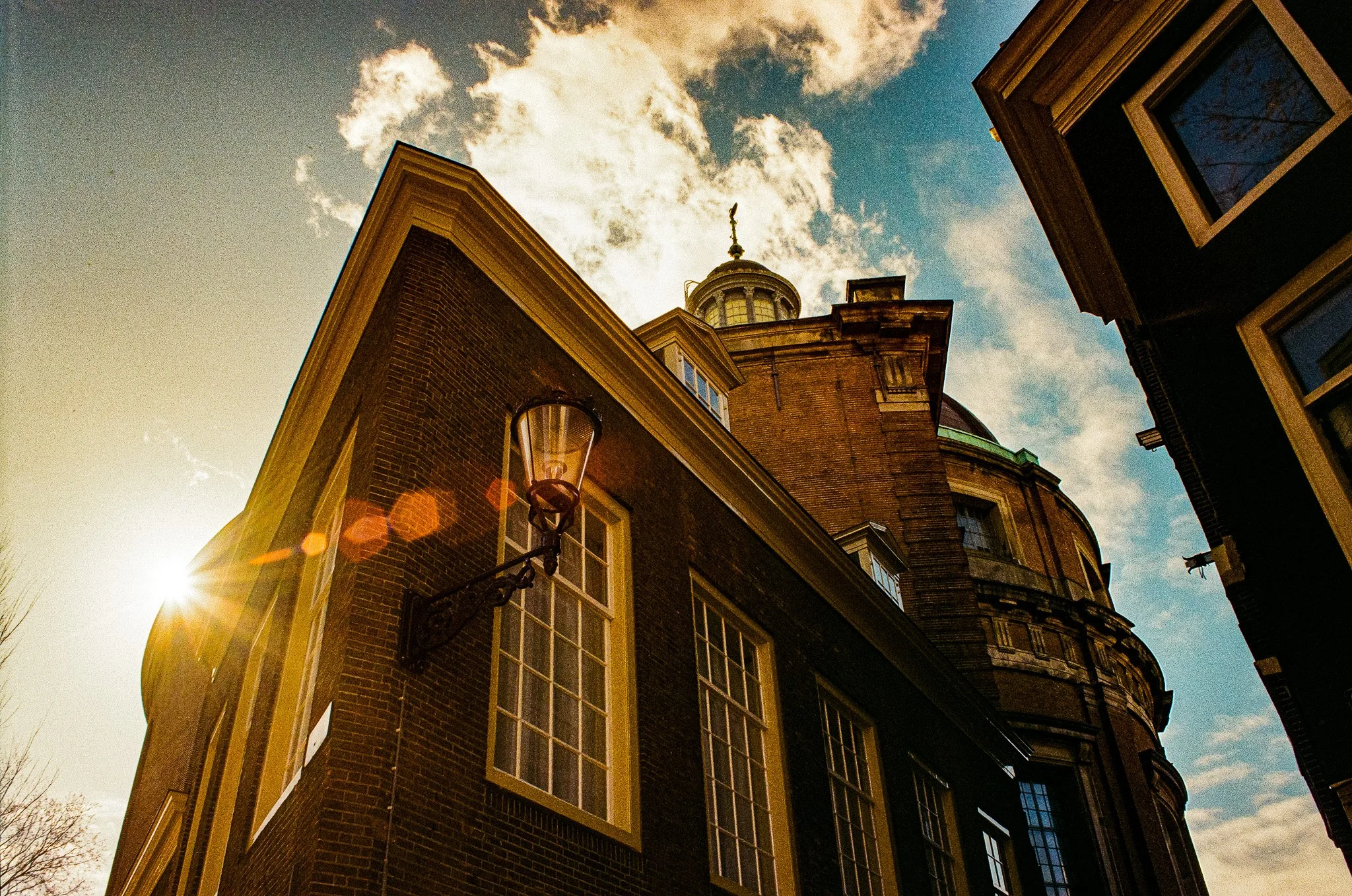 Low-angle view of historic brick buildings with large windows and corner tower, sunlight coming from the left, sky with clouds in background.