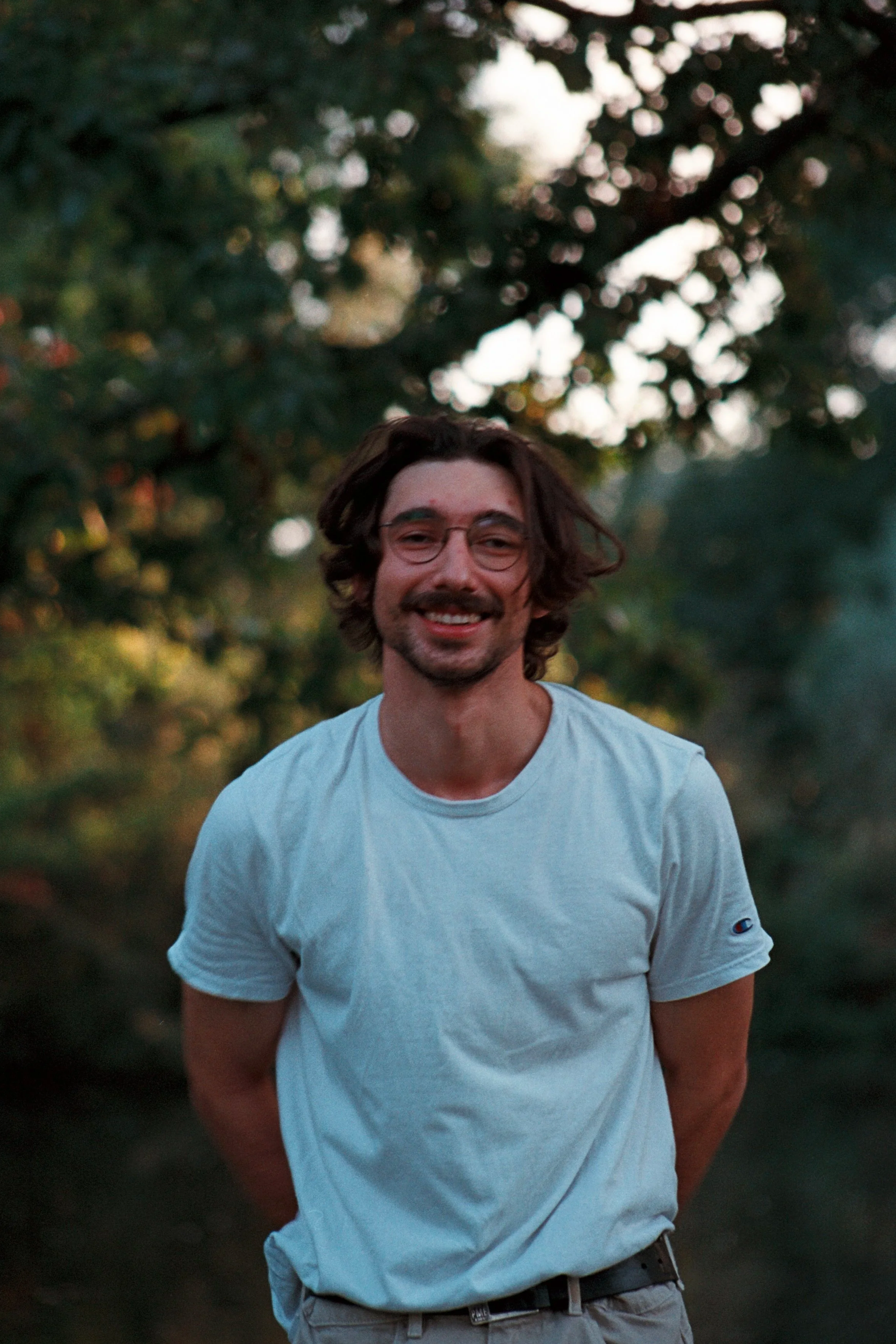A man with glasses, dark wavy hair, and a light beard smiling outdoors in front of trees during sunset, wearing a white T-shirt.