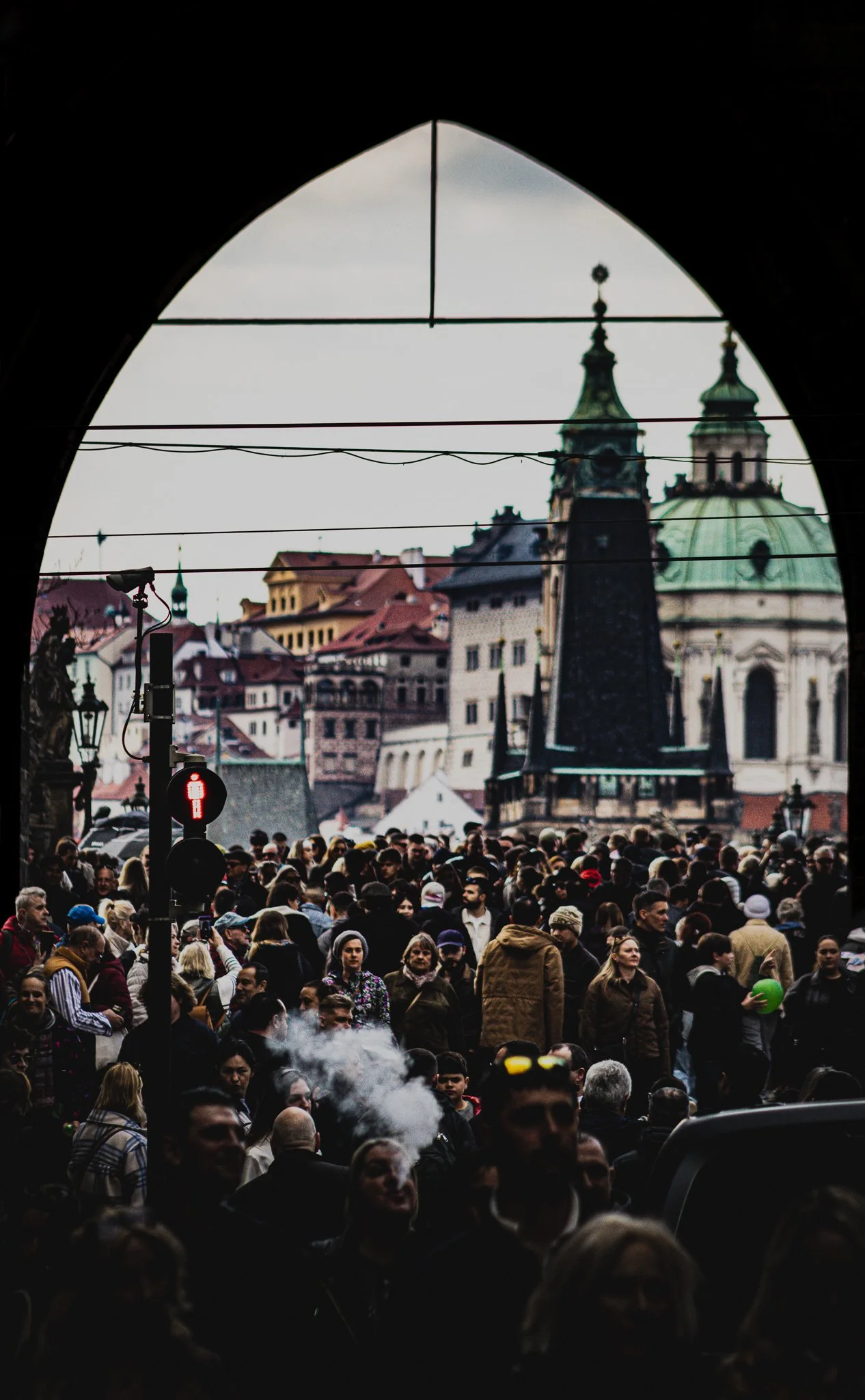 Crowded city street viewed through an archway, with historic buildings and domed towers in the background, and a traffic light showing a red pedestrian signal.