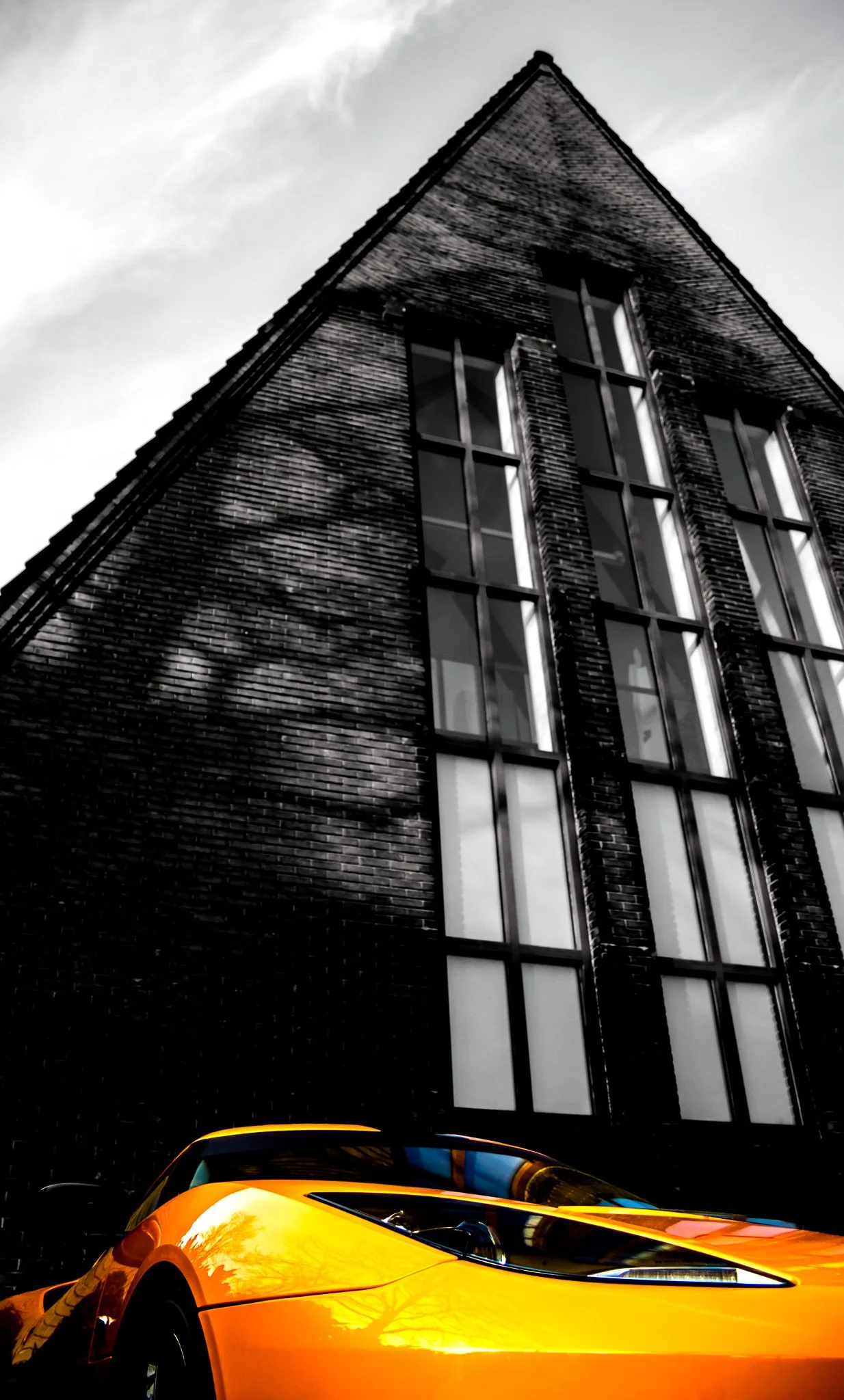 A yellow sports car parked in front of a tall, dark brick building with large windows, captured from a low angle. The sky is partly cloudy.