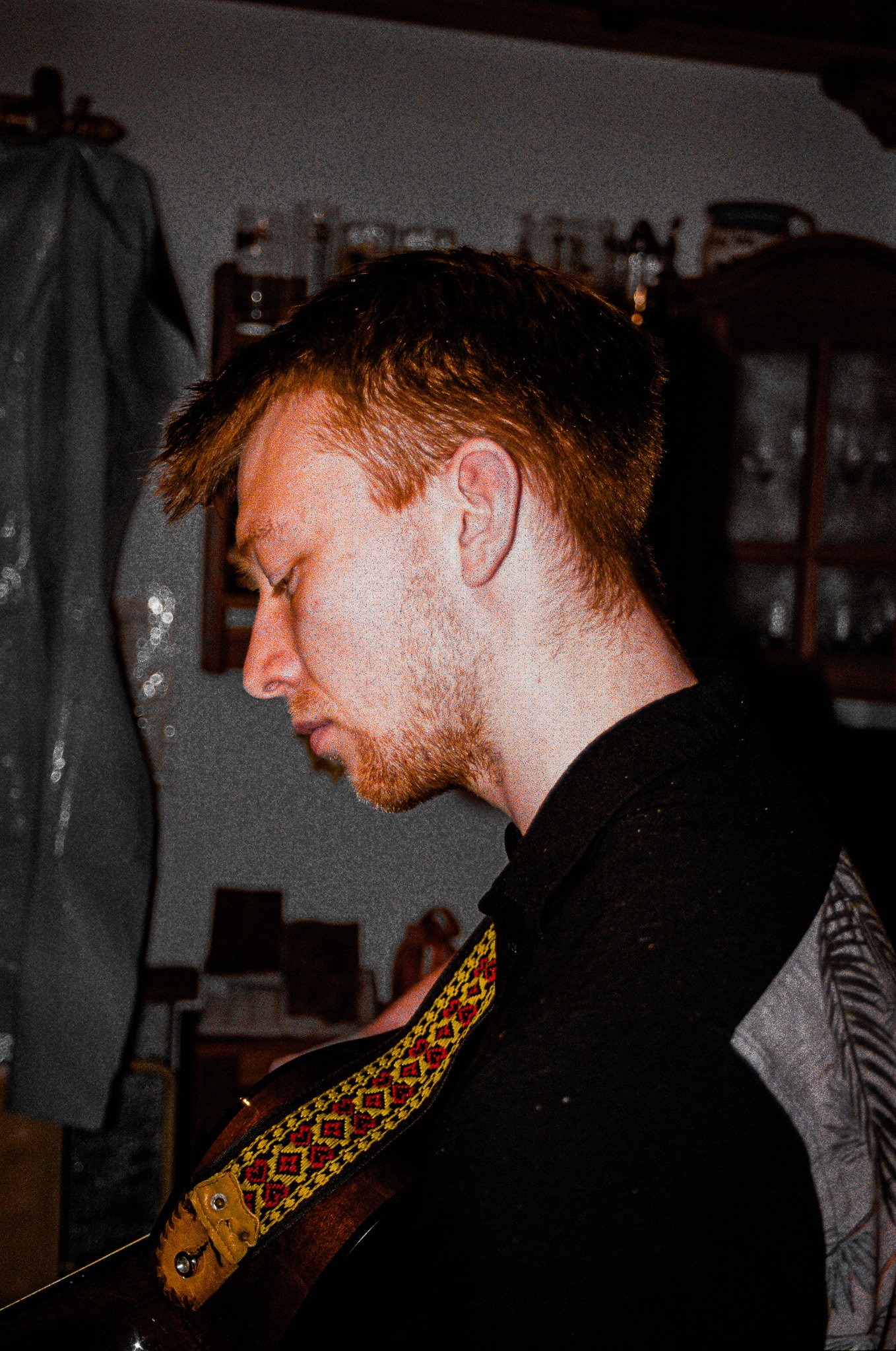 A young man with red hair and a beard, seen in profile, appears to be playing an acoustic guitar with a colorful woven strap, in a room with various objects and furniture in the background.