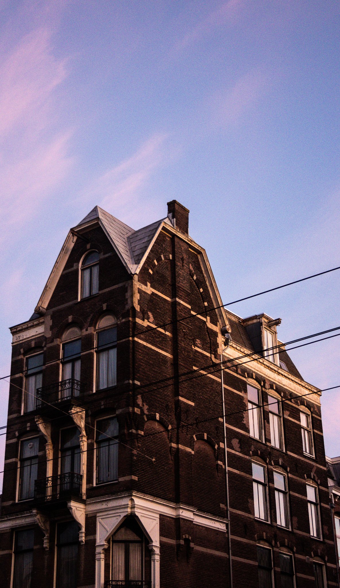 A tall, historic brick building with a peaked roof and multiple ornate windows under a purple sky, with overhead wires crossing in front.