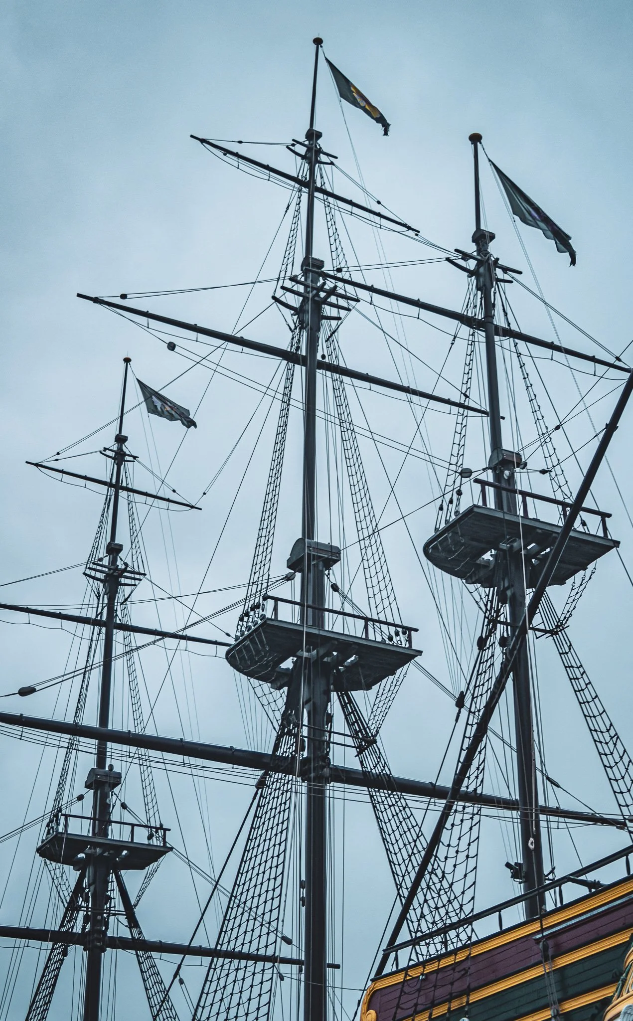 The image shows the masts and rigging of an old sailing ship with flags on top, against a cloudy sky background.