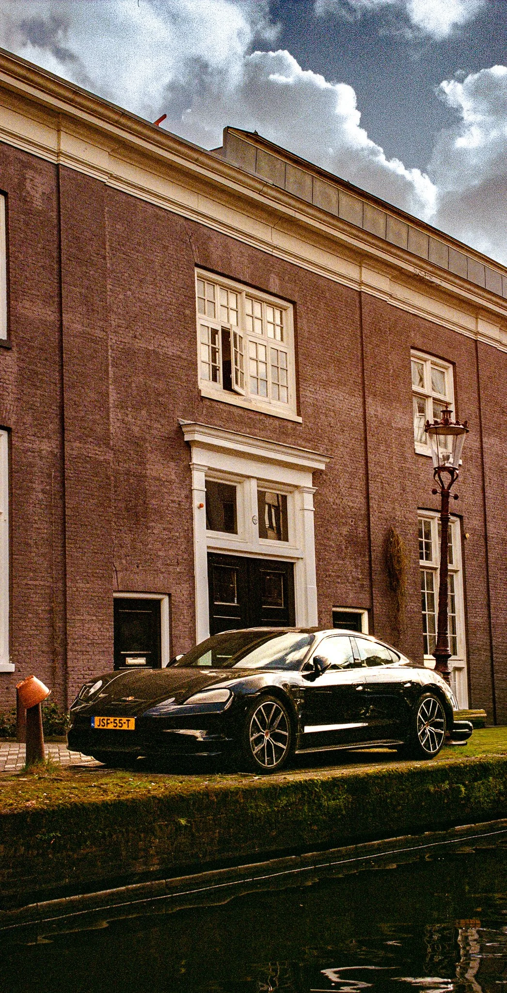A black sports car parked on a grassy area beside a sidewalk in front of a brick building with white trim and multiple windows. A streetlamp is visible near the car, and the sky is partly cloudy.
