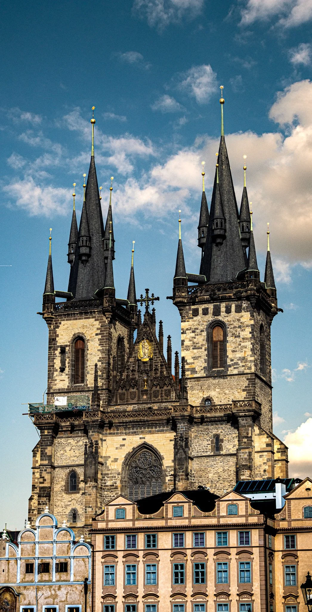 A Gothic-style church with two tall steeples and elaborate architectural details, set against a partly cloudy sky, with surrounding colorful buildings.