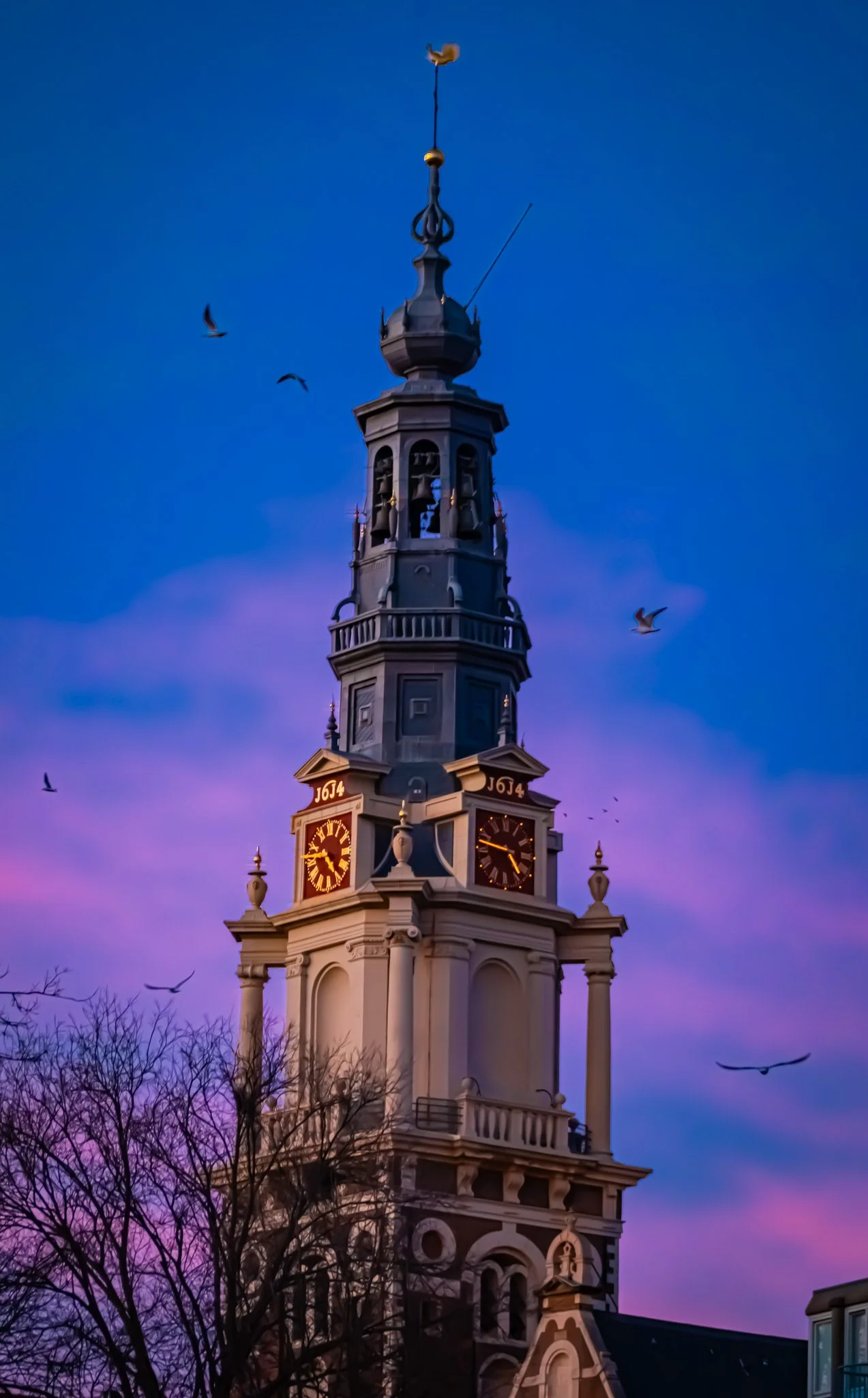 A tall clock tower with a spire, surrounded by flying birds during sunset or twilight with a colorful purple and pink sky in the background.