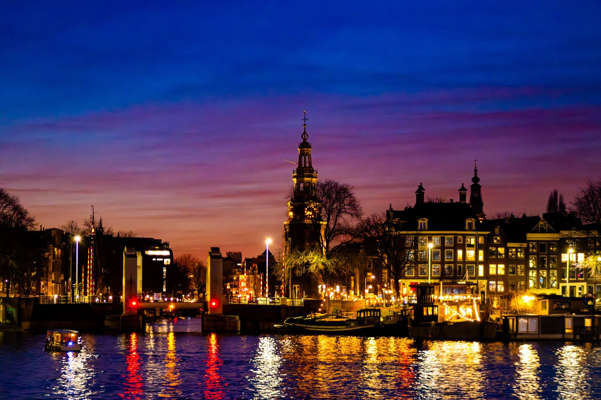Cityscape at dusk with lit buildings, a church tower, and a river reflecting city lights.