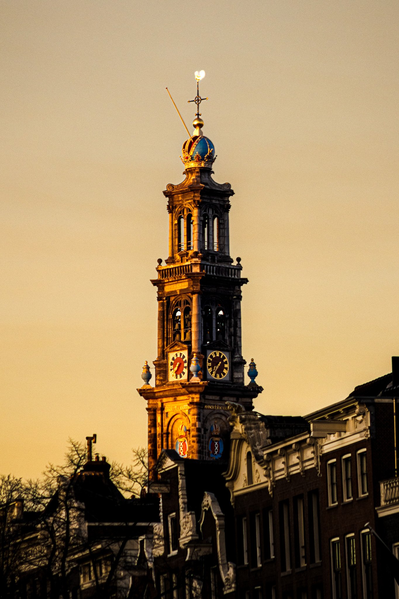 A historic clock tower with a golden crown and weather vane on top, illuminated by warm sunset light, surrounded by old buildings and leafless trees.