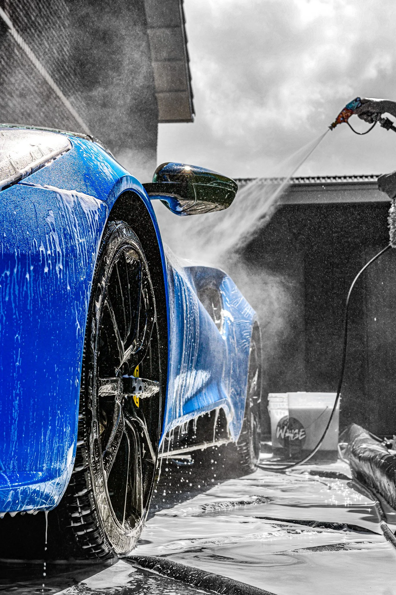 Blue sports car being washed, with water spraying from a pressure washer, on a wet concrete surface.