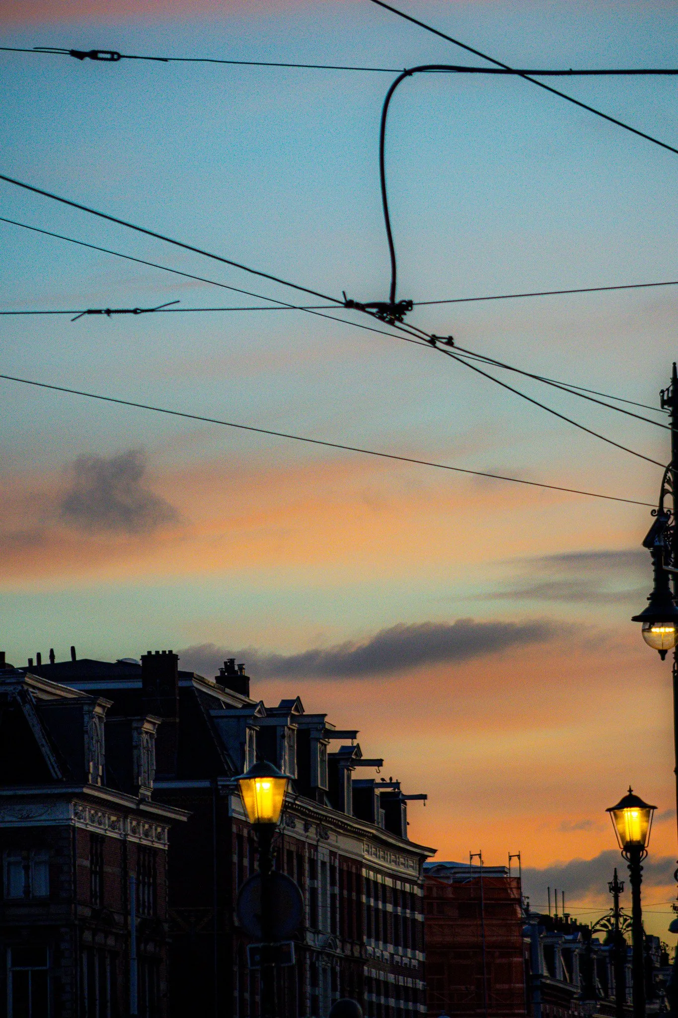 Street view during sunset with illuminated street lamps, historic buildings, and overhead trolley wires against a colorful sky.