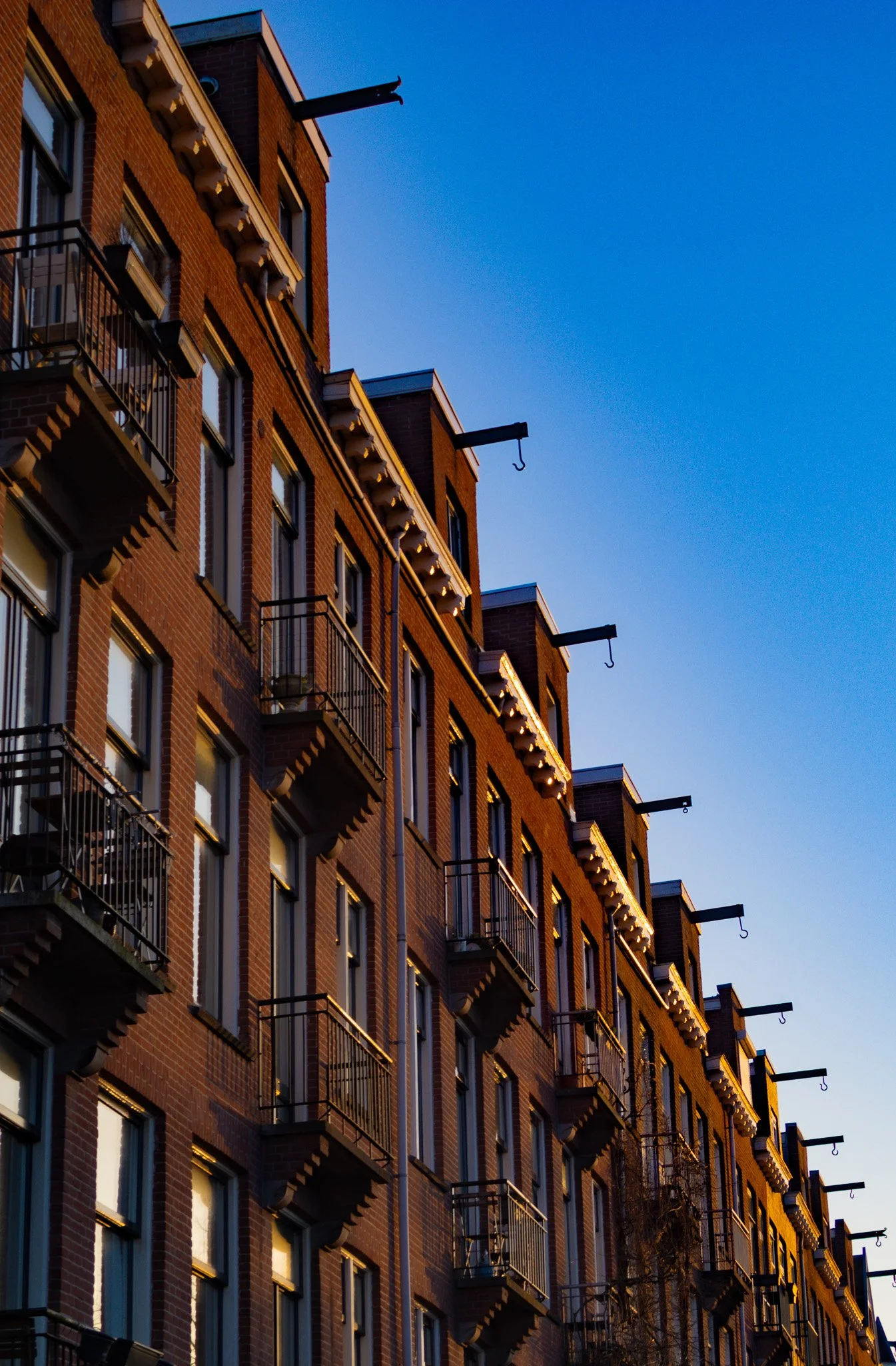 A tall brick apartment building with multiple floors, small balconies with metal railings, and hooks extending from the roof against a clear blue sky.