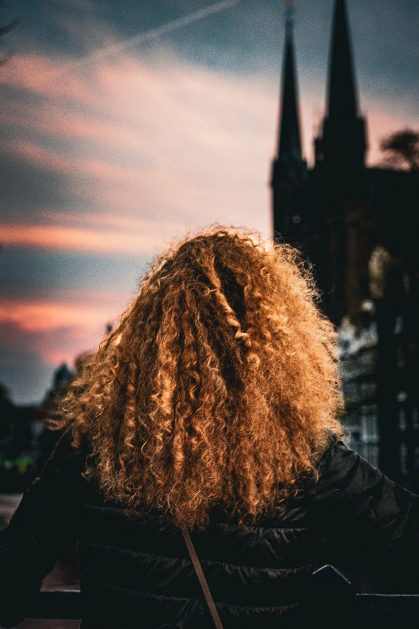 Person with curly hair in a black jacket standing outdoors at sunset with a silhouette of a church with twin spires in the background.
