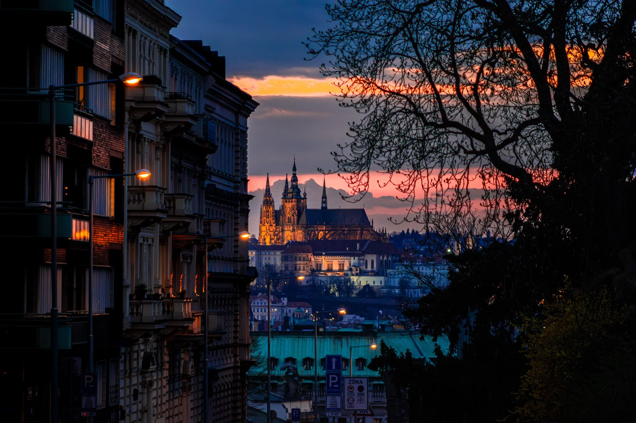 Cityscape at dusk featuring a street with apartment buildings on the left, streetlights turned on, a large cathedral in the background illuminated by sunset, and trees with bare branches in the foreground.