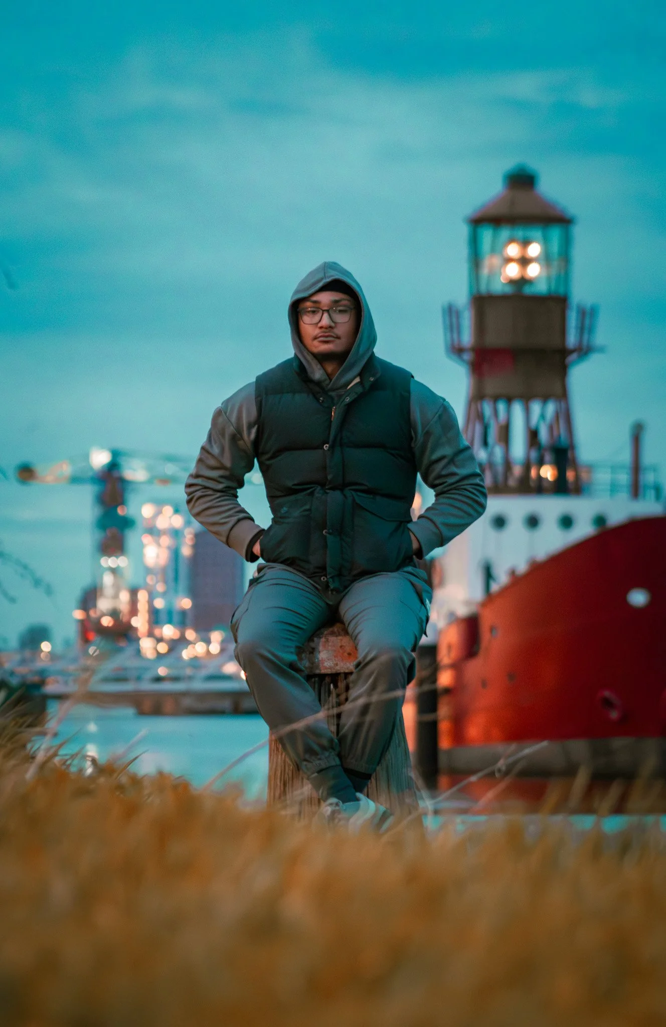 A young man sitting on a wooden post near a lighthouse and boats during dusk, wearing a hoodie, vest, and glasses.