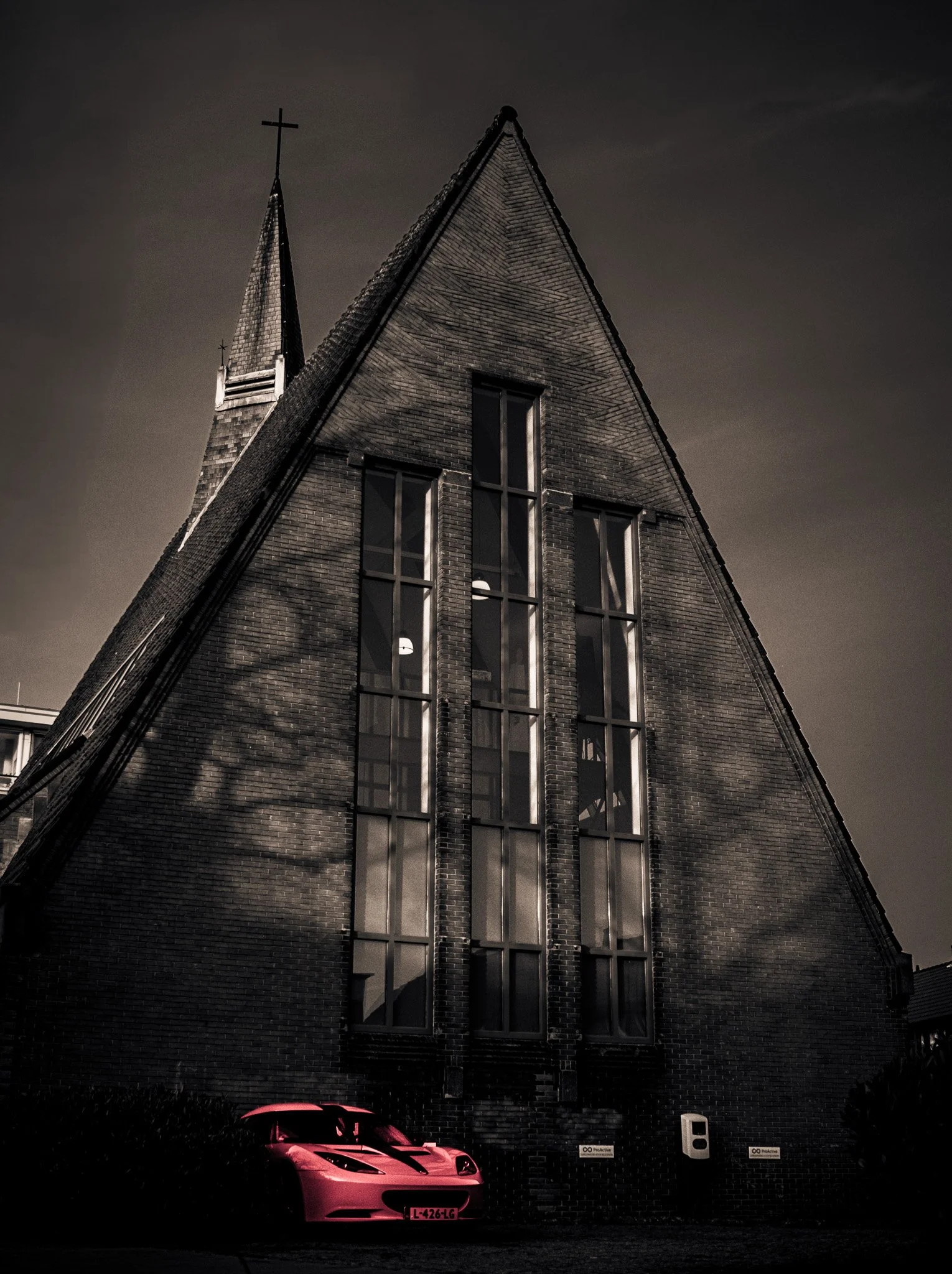 A pink sports car parked in front of a tall brick church with large vertical windows and a steeple with a cross, under a cloudy sky.