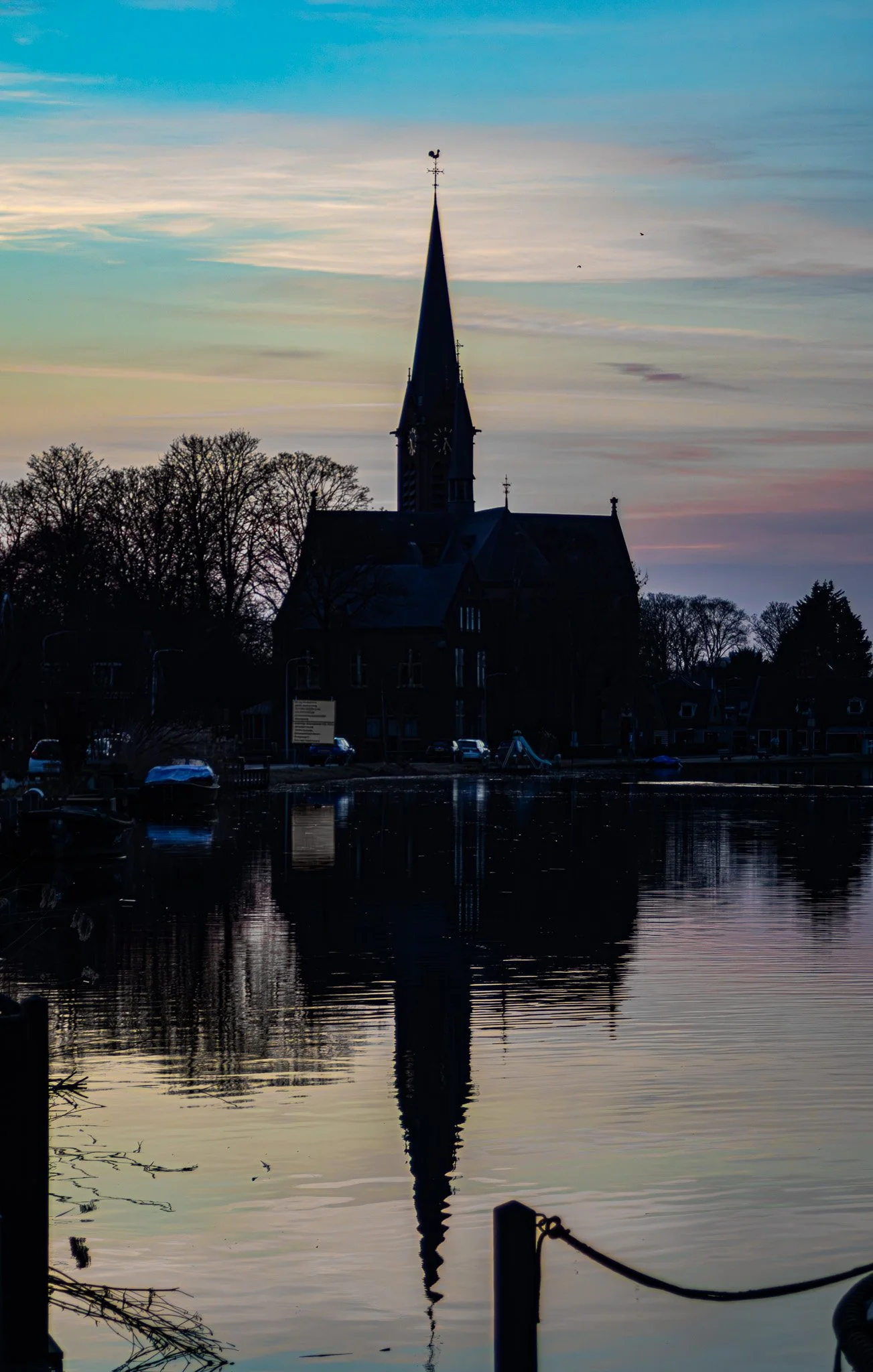A silhouette of a church with a tall steeple reflected in a body of water during sunset or sunrise. The sky features light clouds with pastel colors, and there are trees and parked cars around the water.