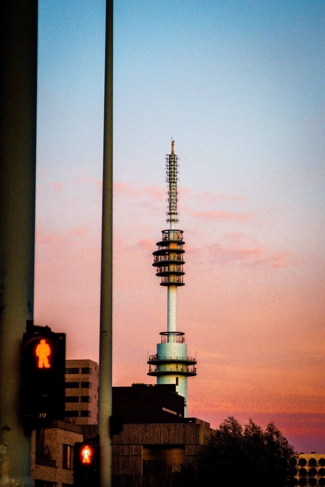 City skyline at sunset with a tall communication tower and red pedestrian crossing signals in the foreground.