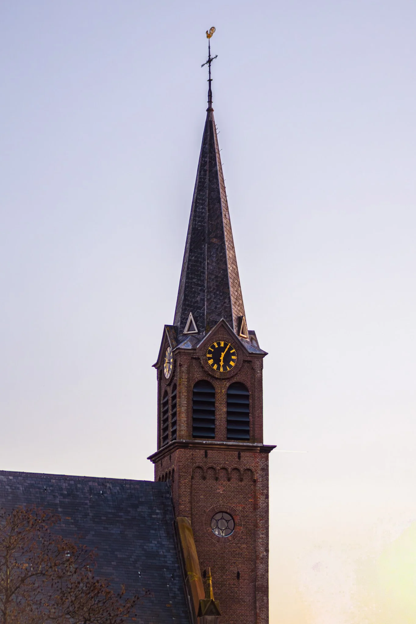 A church steeple made of brick with a clock face, topped by a pointed dark shingled roof and a weather vane.