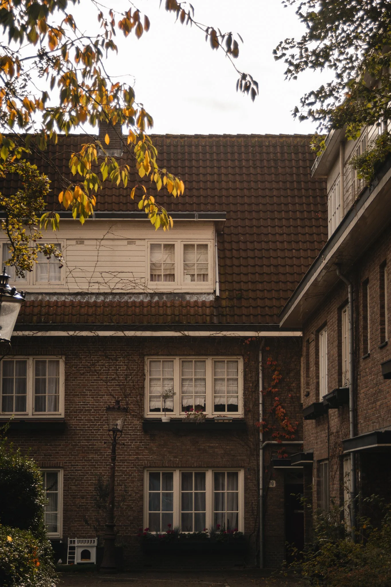 View of a traditional brick house with multiple windows, a brown tiled roof, and nearby trees with yellow and green leaves.