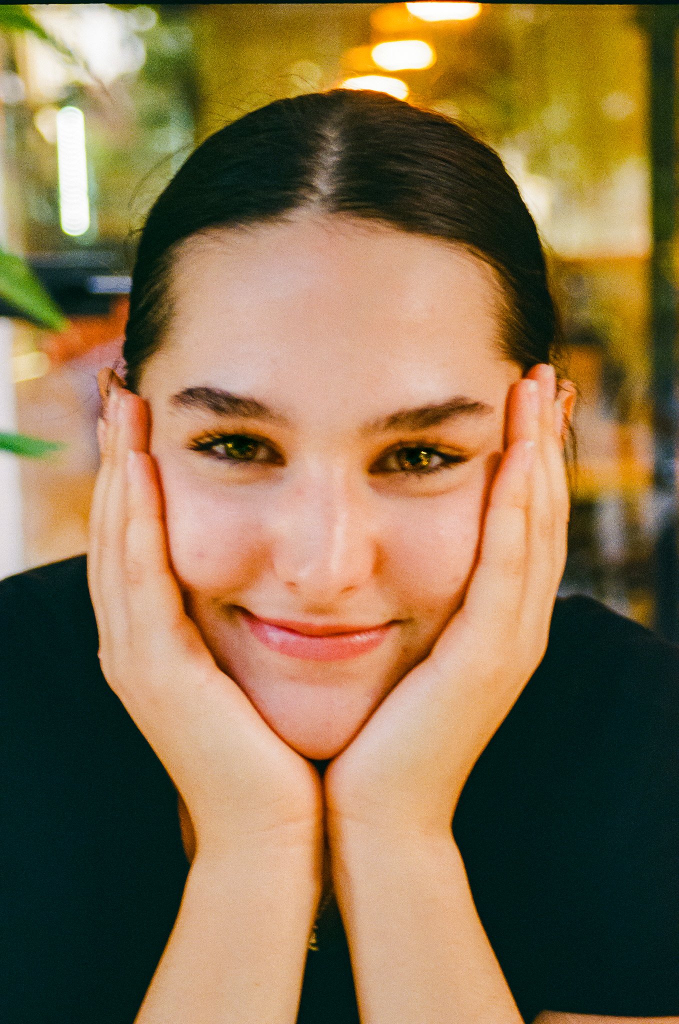 Close-up of a smiling woman with dark hair, resting her face on her hands in a lively indoor setting with warm lighting and blurred background.