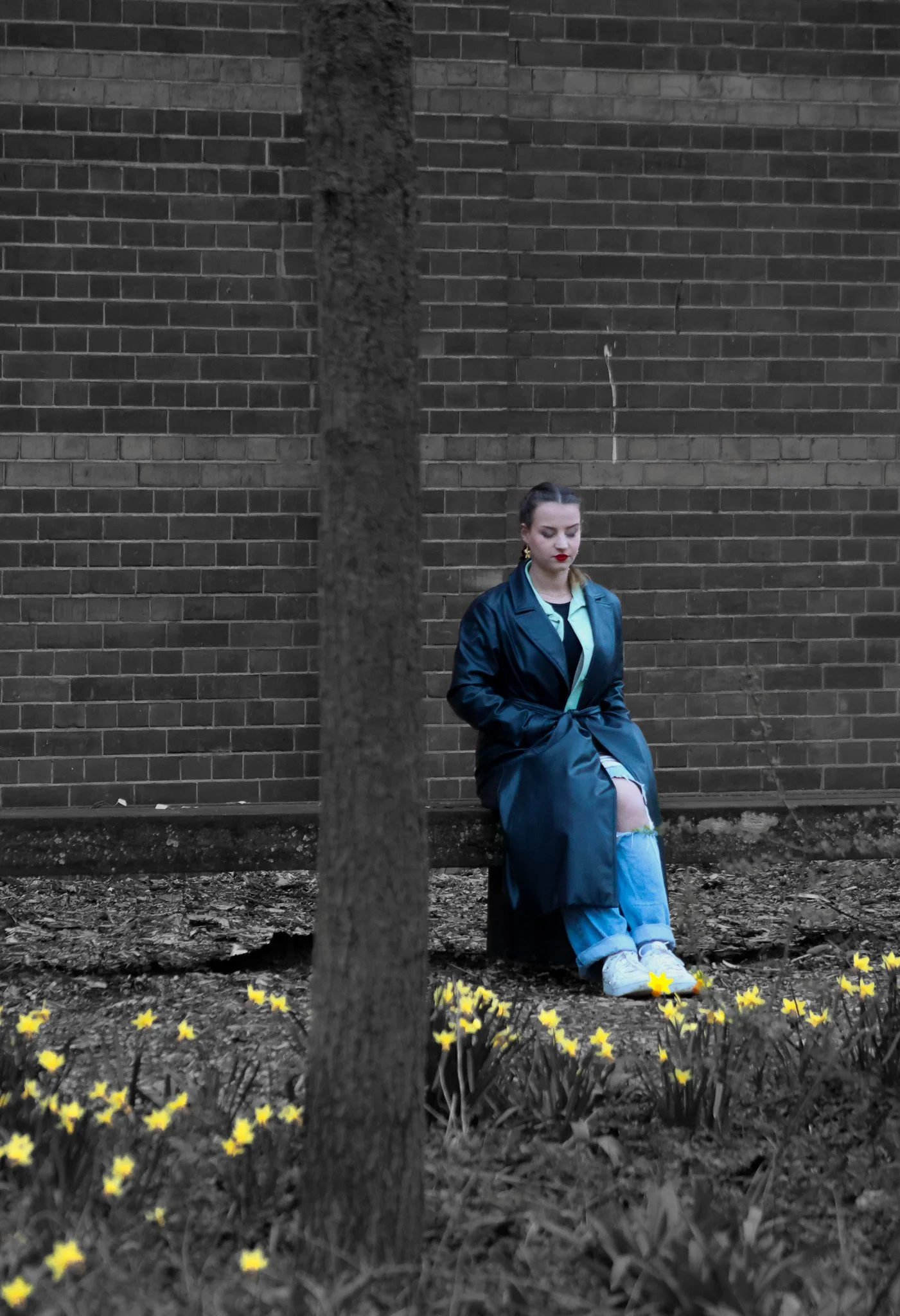 A young woman sitting on a wooden bench with her head bowed, wearing a black raincoat and ripped jeans, in front of a brick wall with yellow flowers in the foreground and a tree partially obscuring her.