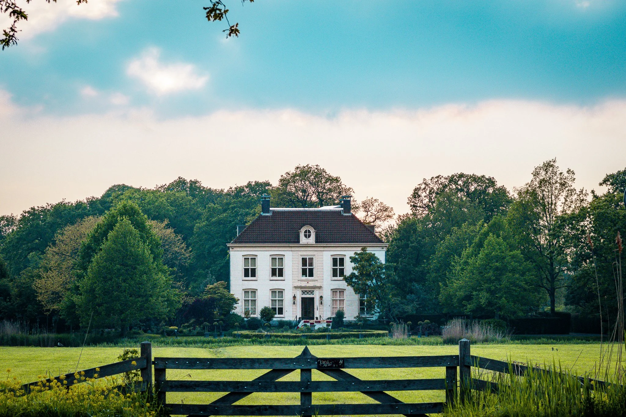A large white house with multiple stories and an ornate front door, surrounded by green trees, with a wooden fence and a green lawn in the foreground, under a partly cloudy sky.