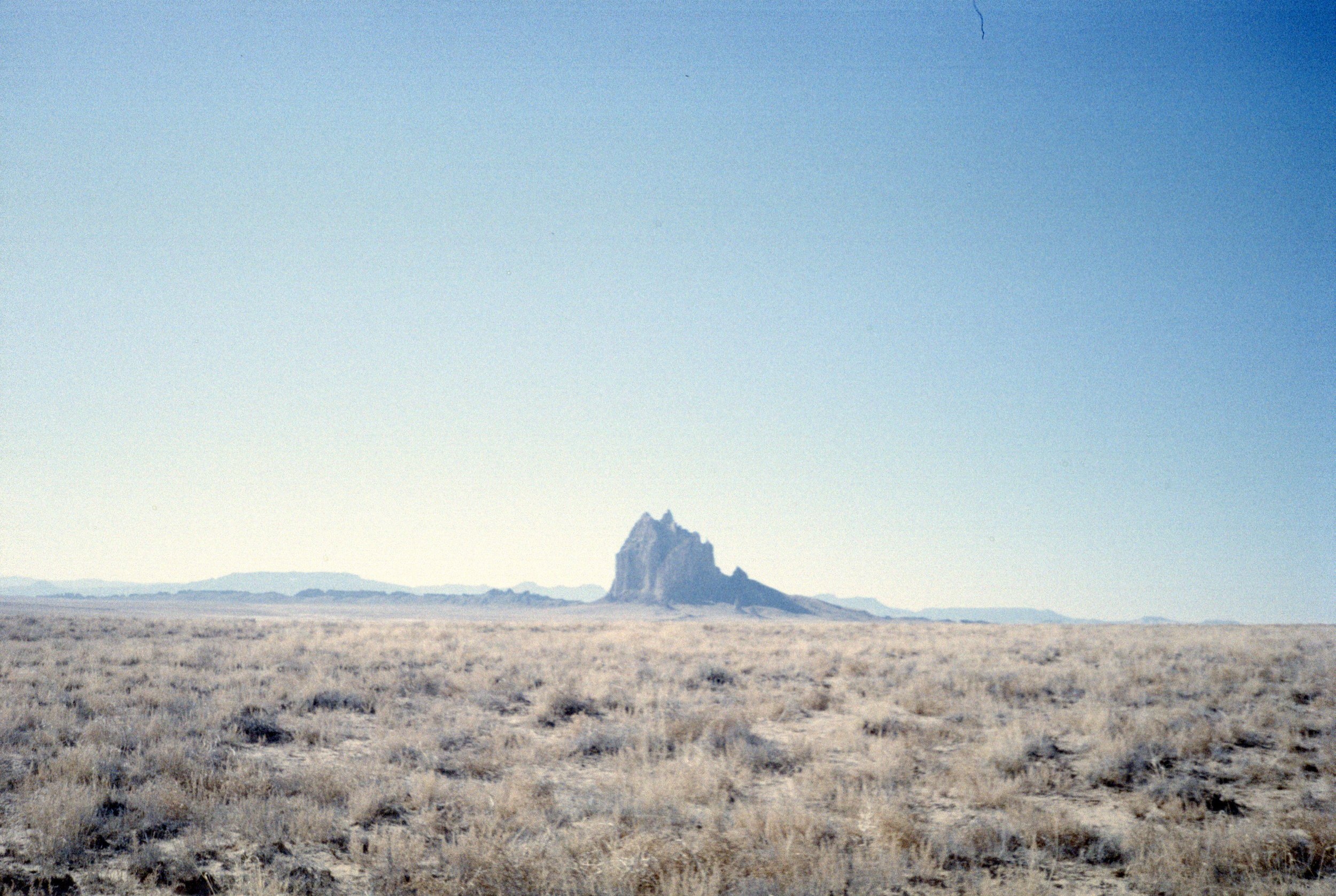 A vast, flat desert landscape under a clear blue sky, with a prominent jagged rock formation in the distance.