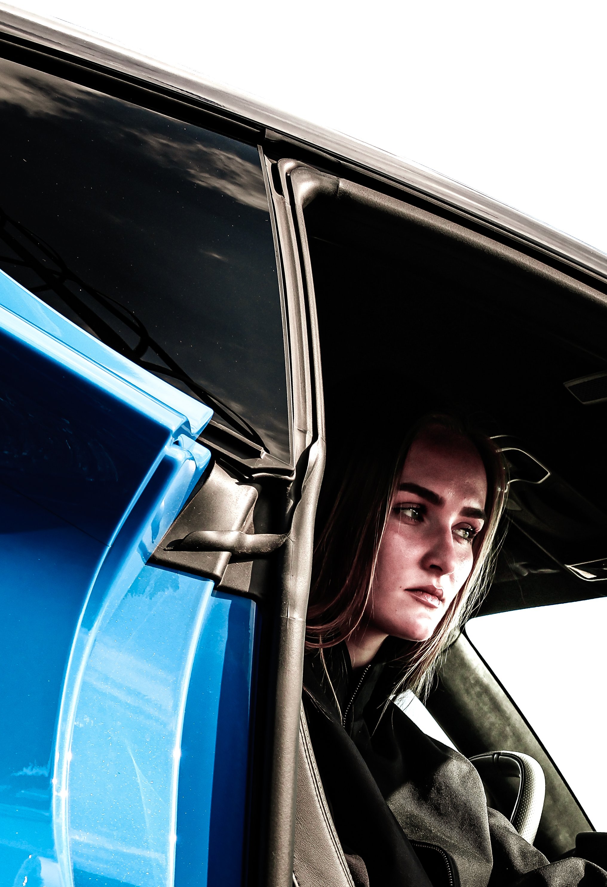 A woman with long brown hair sitting in a vehicle with a black interior. She appears to be looking ahead and slightly to the side, wearing a black top or jacket. The photo is taken from outside the vehicle, focusing on her face and part of the driver