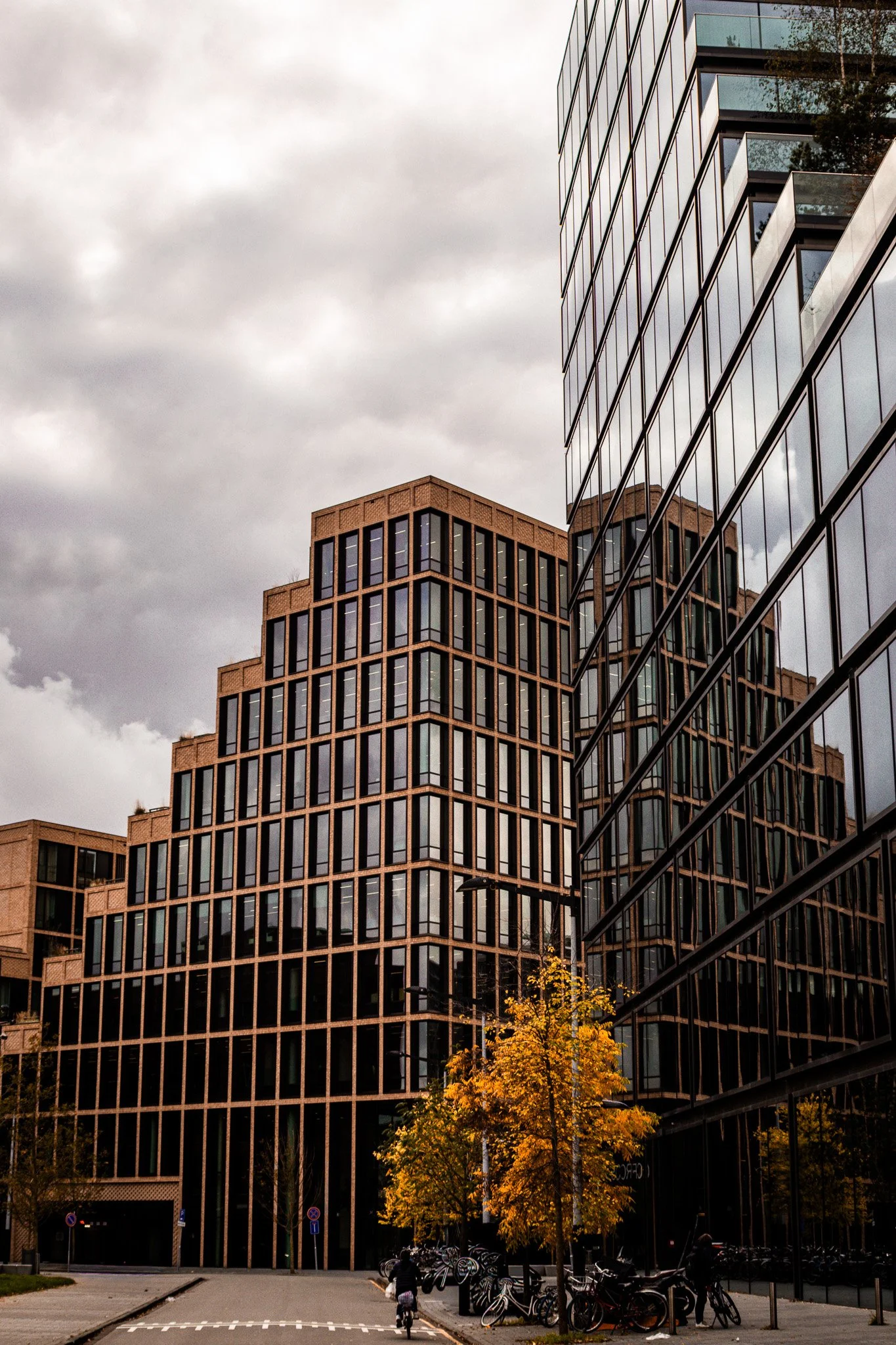 Modern office buildings with large glass windows, one reflecting another building, against a cloudy sky and autumn trees with yellow leaves.