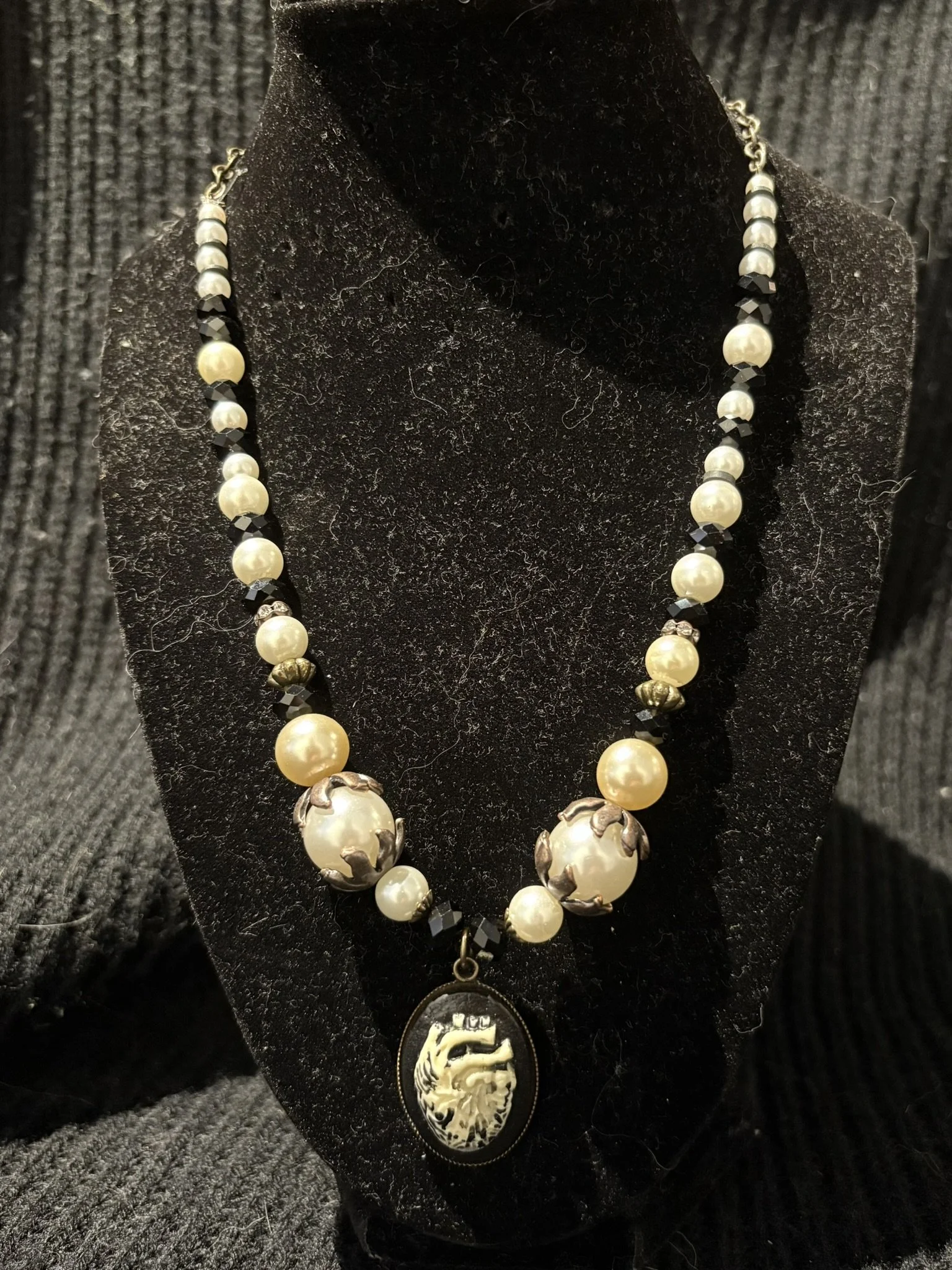 A necklace featuring large cream-colored pearls and small black beads with silver decorative elements, hanging from a black velvet display bust.
