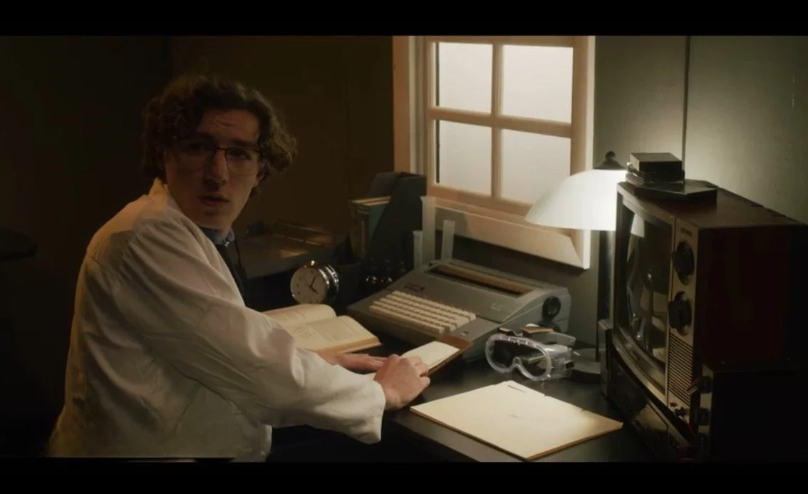 Man with glasses sitting at a cluttered desk with vintage computer, radio, and books in a dimly lit room.