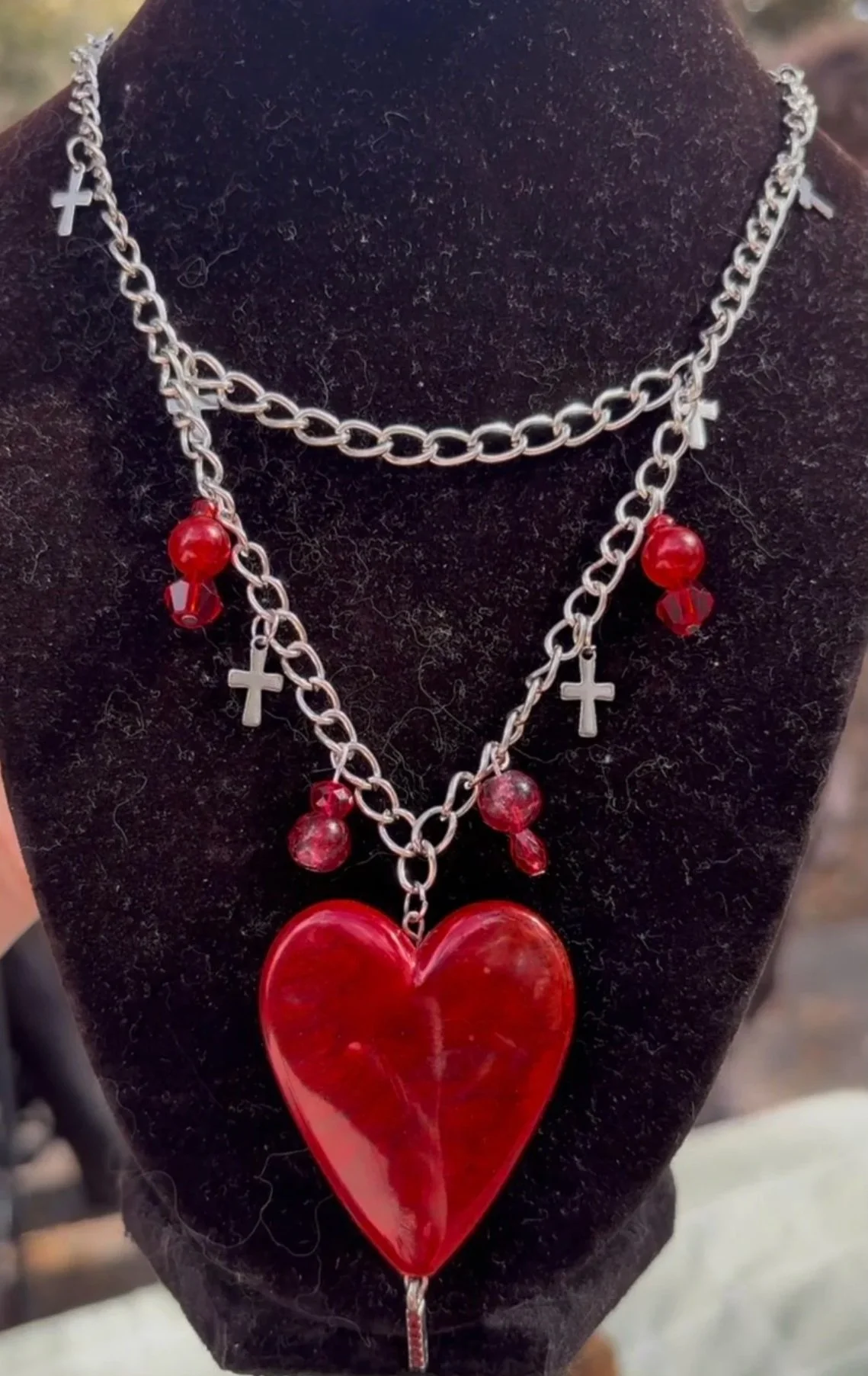 A black display bust wearing layered silver chains with religious crosses and red heart-shaped pendant, accented with red beads.